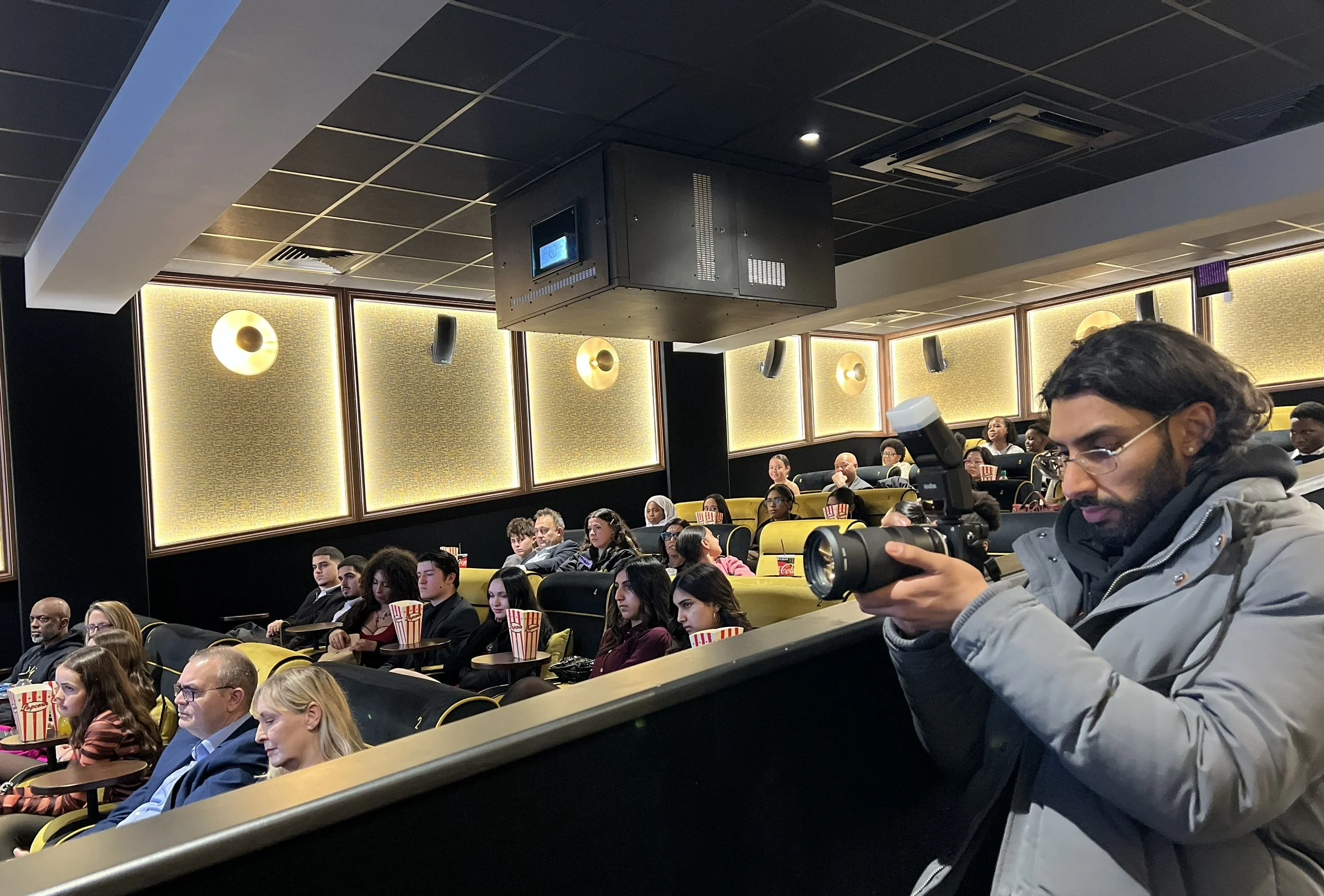 A man with glasses and a gray jacket uses a camera to take photos while seating in a theater or cinema filled with people watching a presentation.