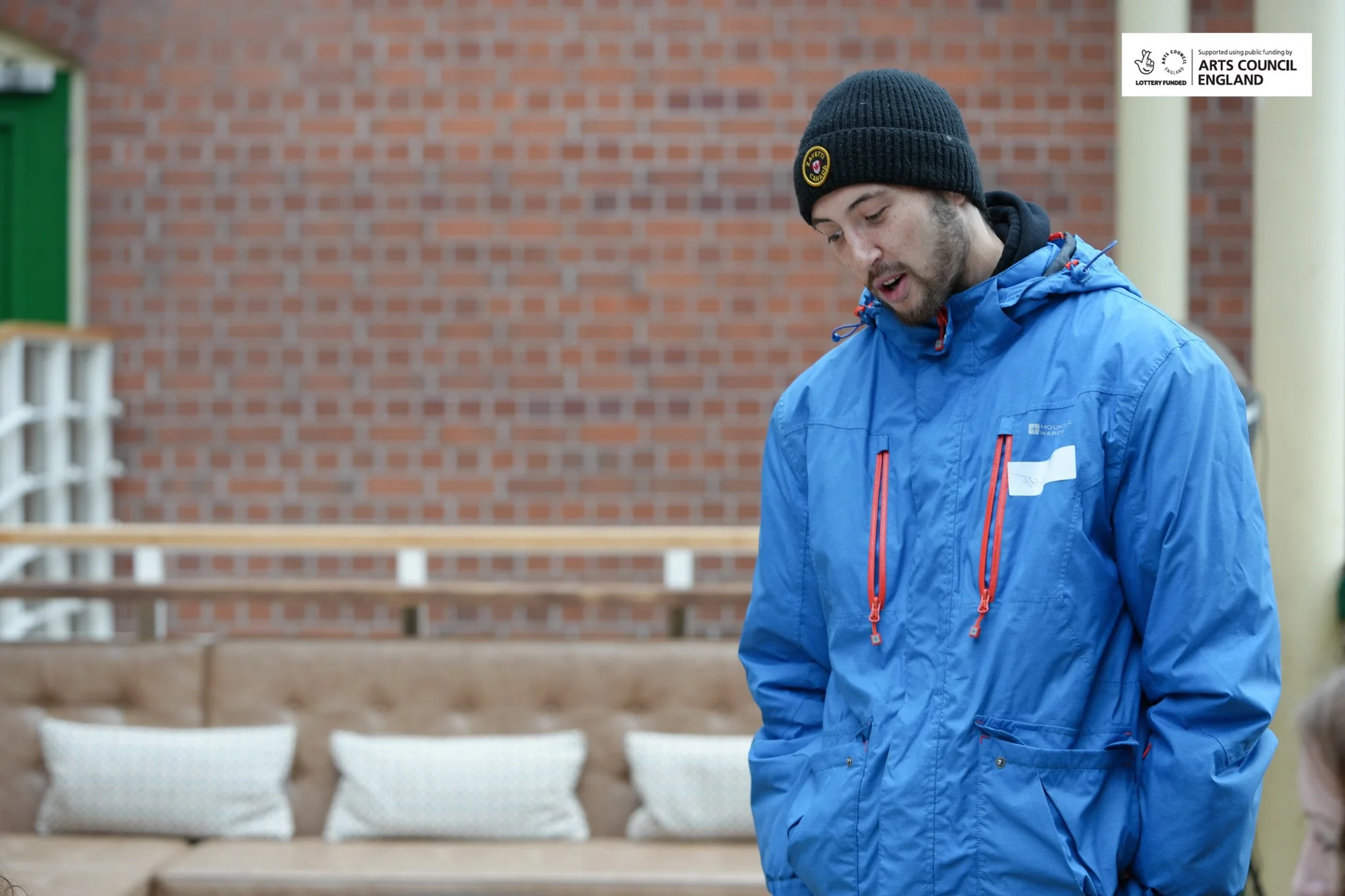 A man in a blue jacket and a black knit cap looks down, standing in front of a brick wall and a beige sofa with pillows.