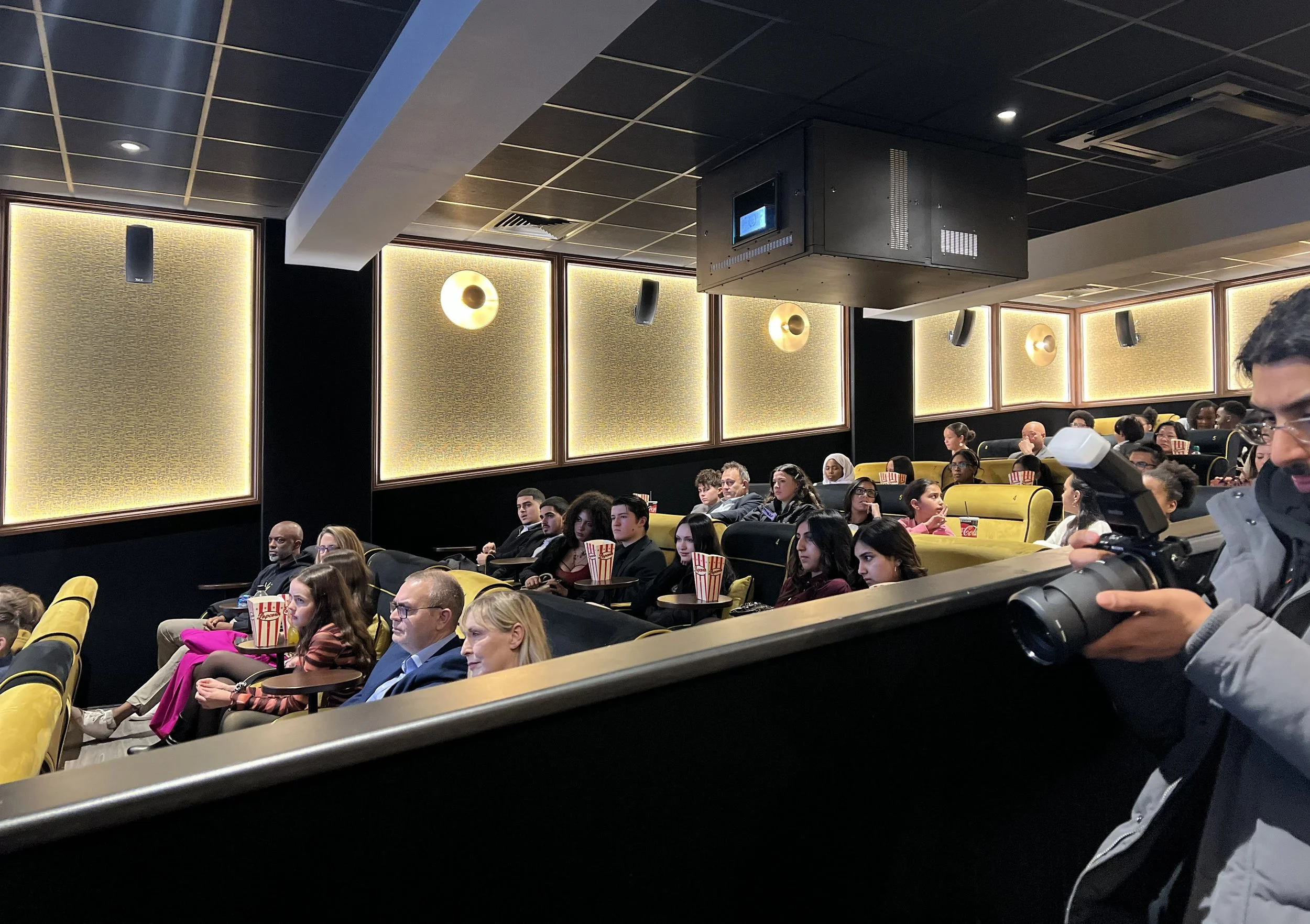 Audience watching a presentation in a theater with tiered yellow seats and movie snacks, with a person capturing the event with a camera.