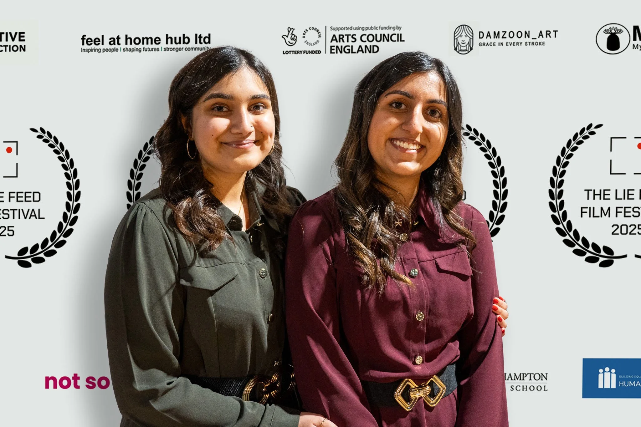 Two women standing together, smiling, in front of a backdrop with film festival and arts logos, wearing colored button-down shirts and belts.