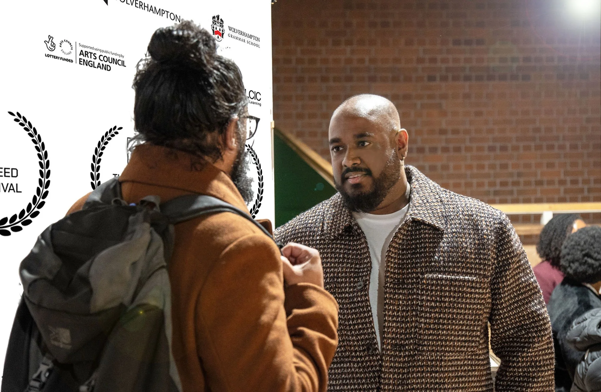 Two men engaged in conversation indoors, with a brick wall and a white banner with logos behind them. One man is facing away with a backpack, the other is facing towards the camera.