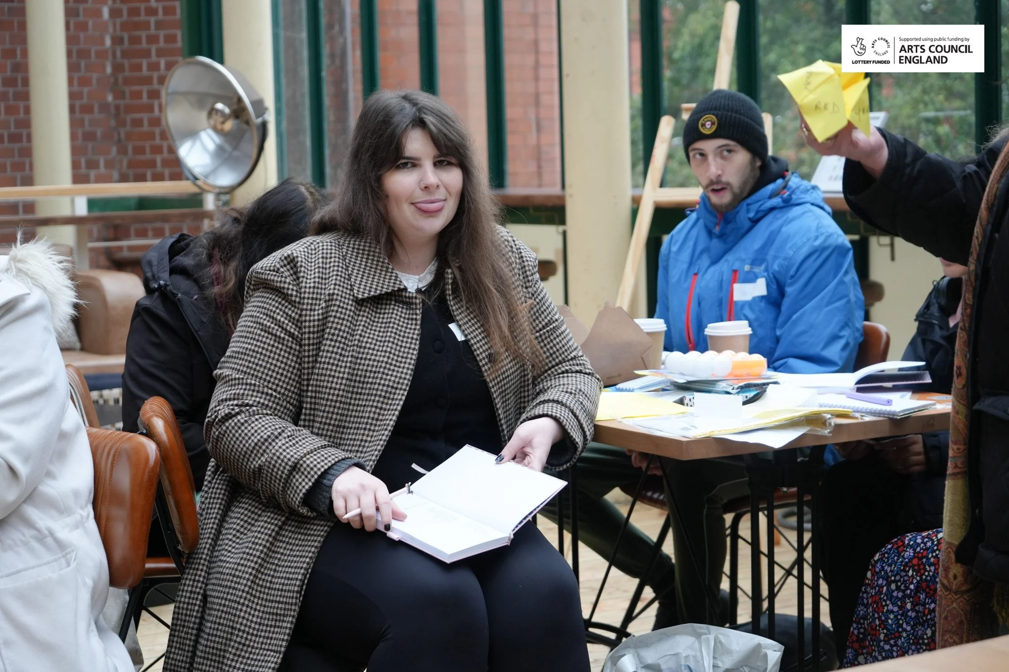 A young woman with long brown hair sticking out her tongue, holding a notebook, sitting at a table with a group of people in a bright indoor setting with large windows, brick wall, and plants.