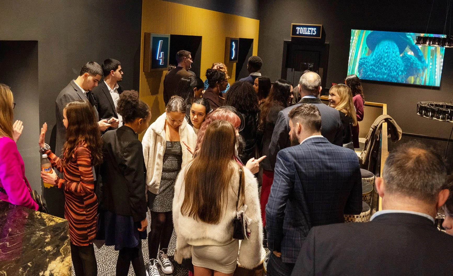 Group of people waiting in line at a venue, with signs for restrooms and a TV screen in the background.