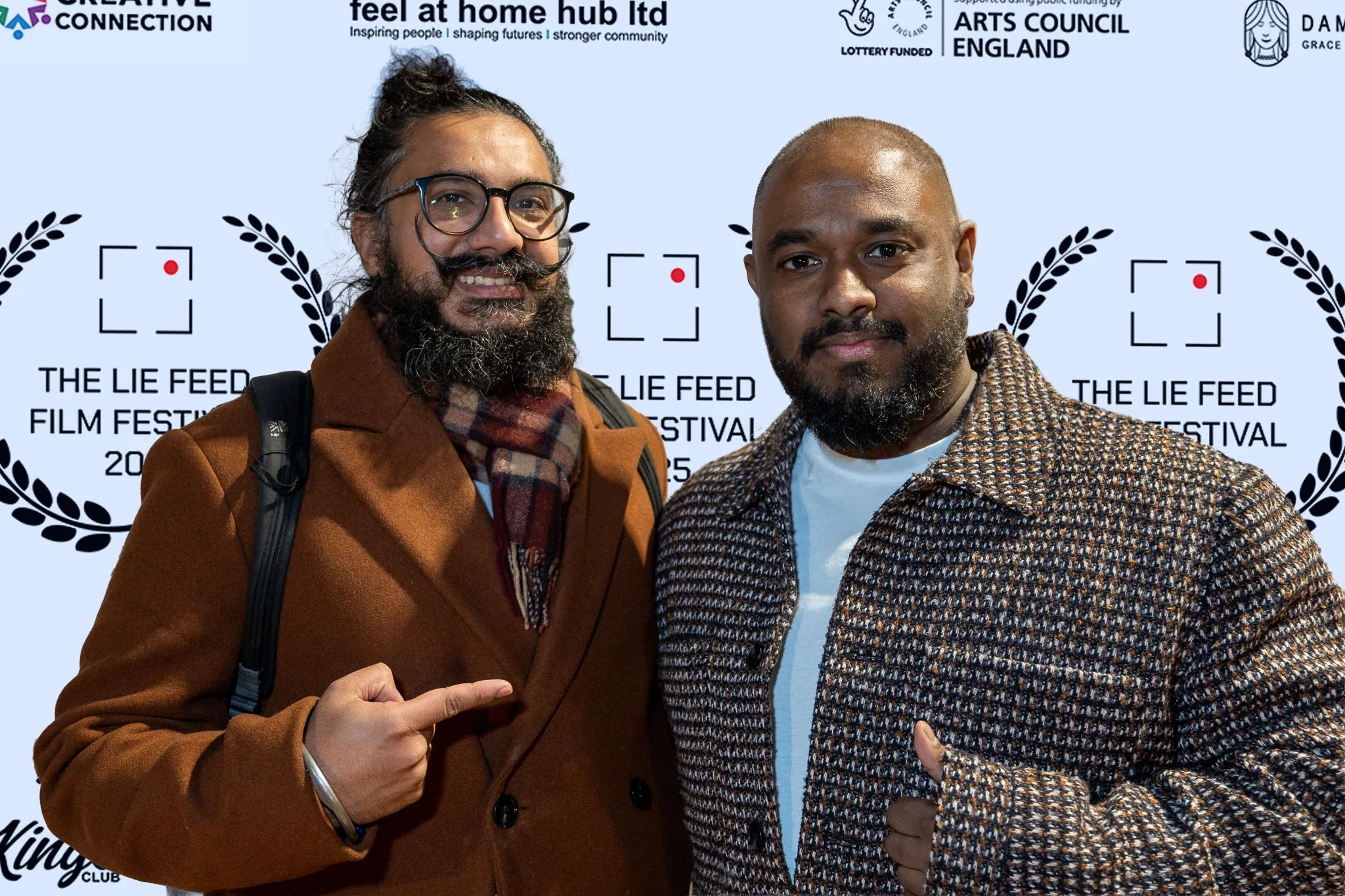 Two men attending a film festival, standing in front of a backdrop with logos and the text 'The Lie Feed Film Festival 2023.' One man has glasses, a beard, and long hair tied back, wearing a brown coat and scarf, pointing at the other man. The second
