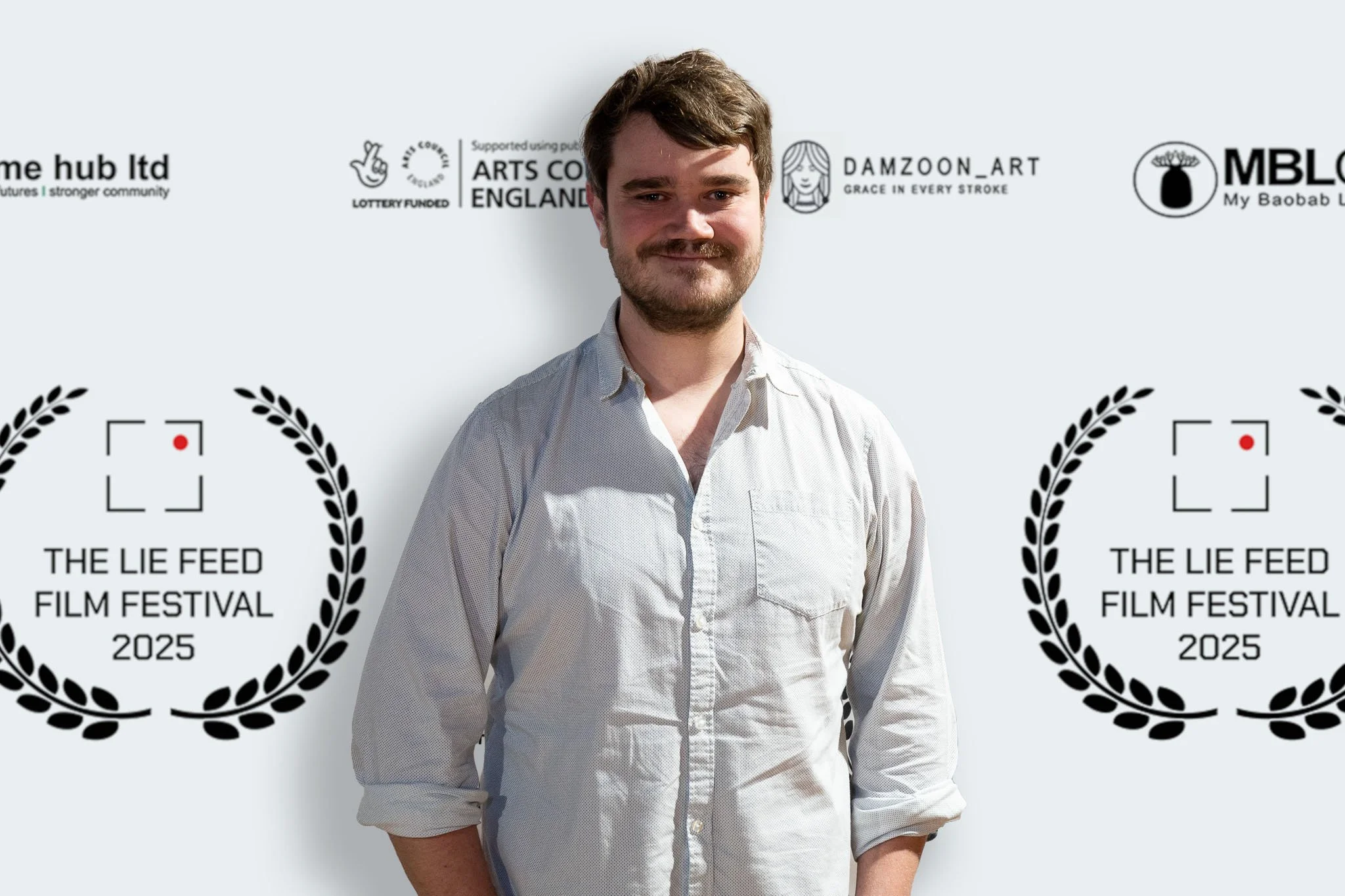 A man with light brown hair and a beard wearing a light beige button-up shirt, standing in front of a white backdrop with logos and text for The Lie Feed Film Festival 2025.