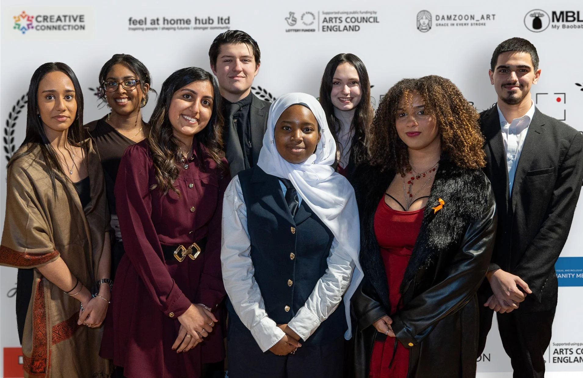 Group of nine diverse young adults standing in front of a white backdrop with logos, dressed in formal and traditional attire, smiling for a photo.