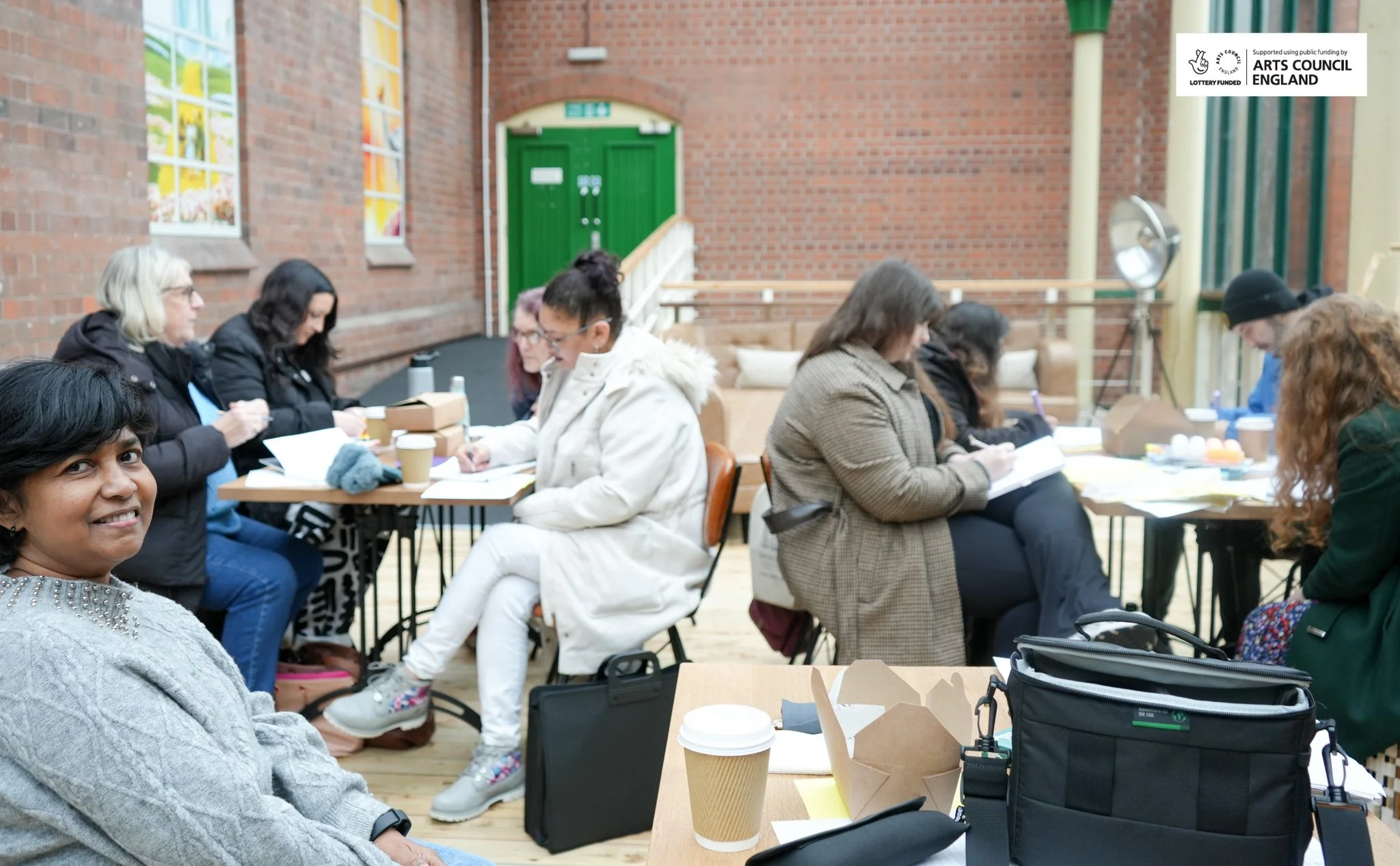 People sitting around tables with notebooks and coffee cups inside a brick-walled room with colorful stained glass windows and a green door.