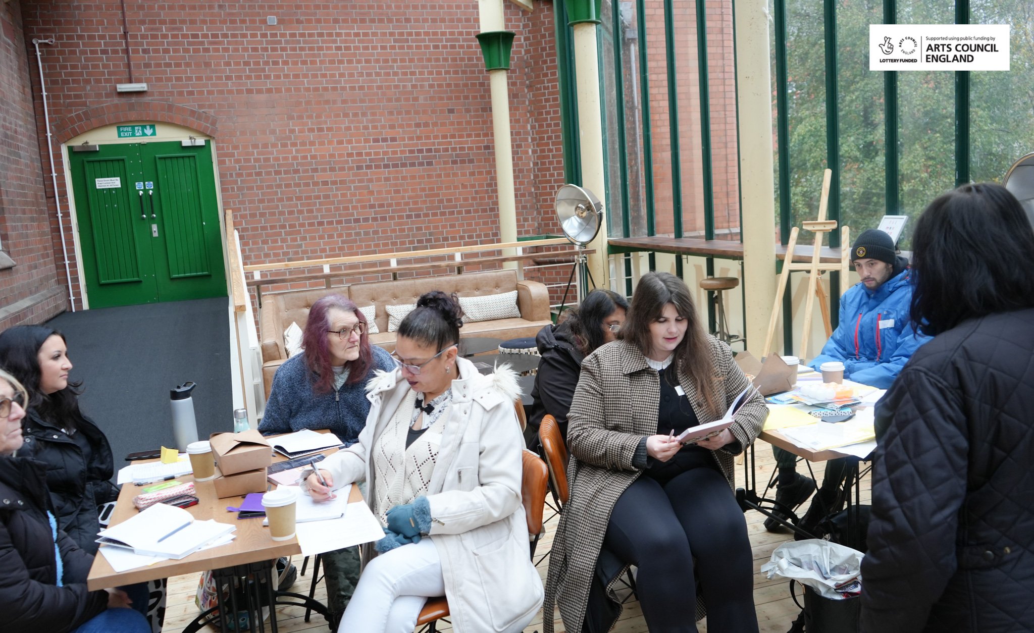 People sitting at tables in a workshop or meeting space with brick walls and large windows, some reading or writing, and a woman standing.
