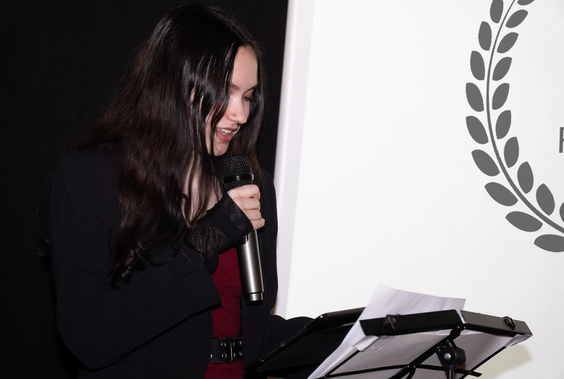 Young woman with long dark hair reading from a paper, holding a microphone, standing in front of a white wall with gray laurel wreath design.