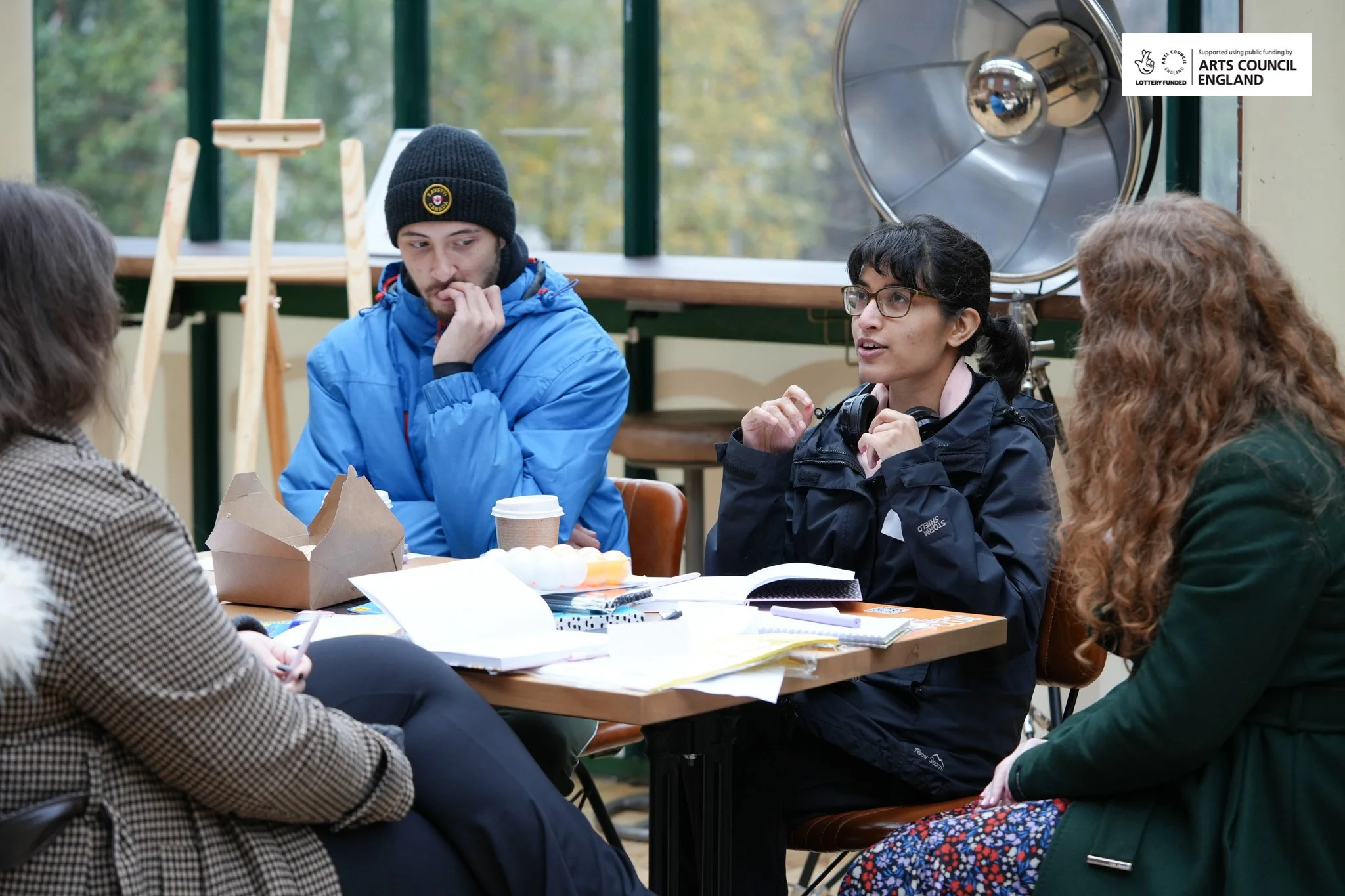Four people sitting around a table in a room with large windows, having a discussion. On the table are notebooks, papers, a coffee cup, and a carton of eggs. One woman is speaking, and the others are listening attentively.