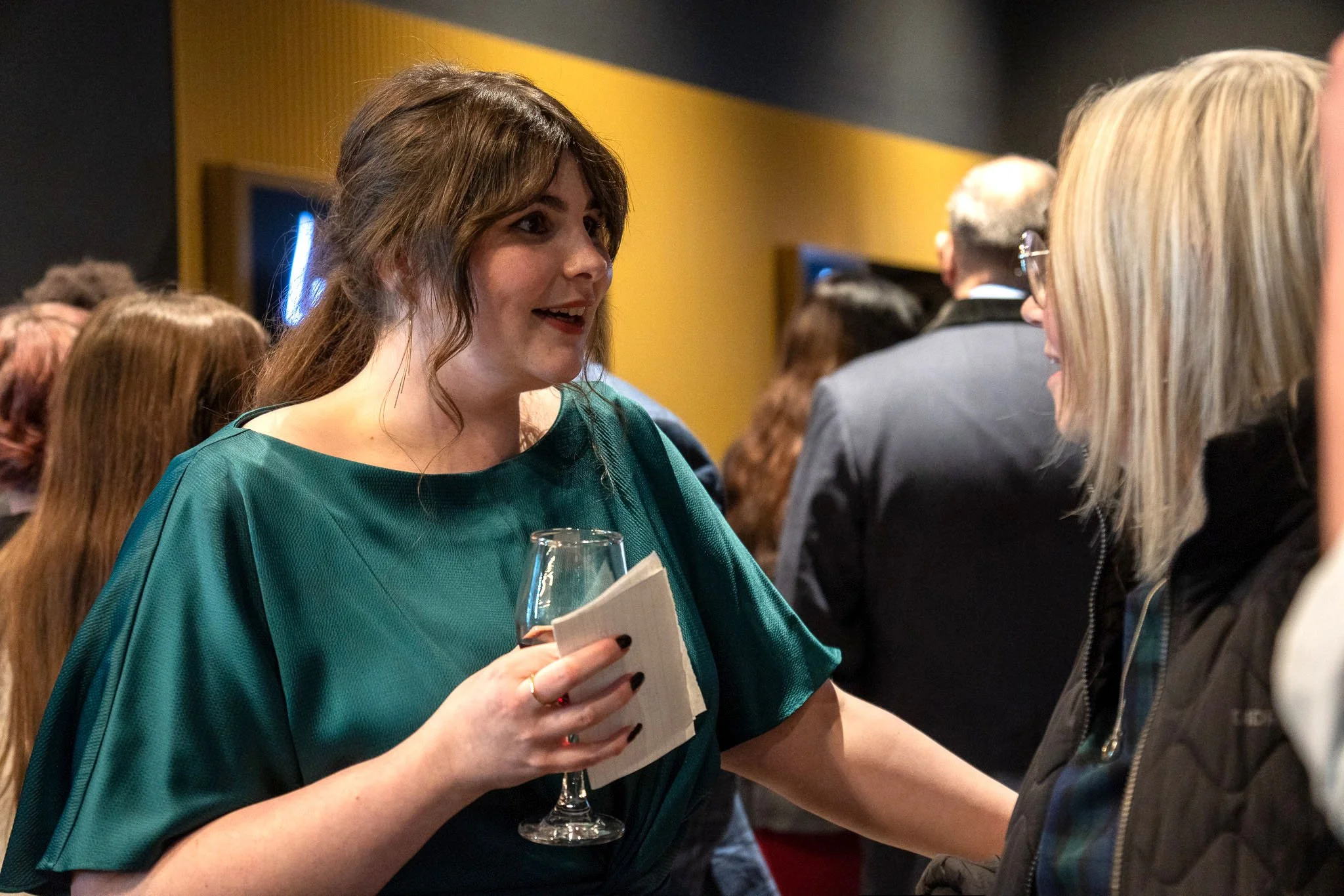Two women are having a conversation at a social event. The woman on the left is holding a wine glass and a paper napkin, while the woman on the right is wearing glasses and a jacket. Several people are visible in the background.