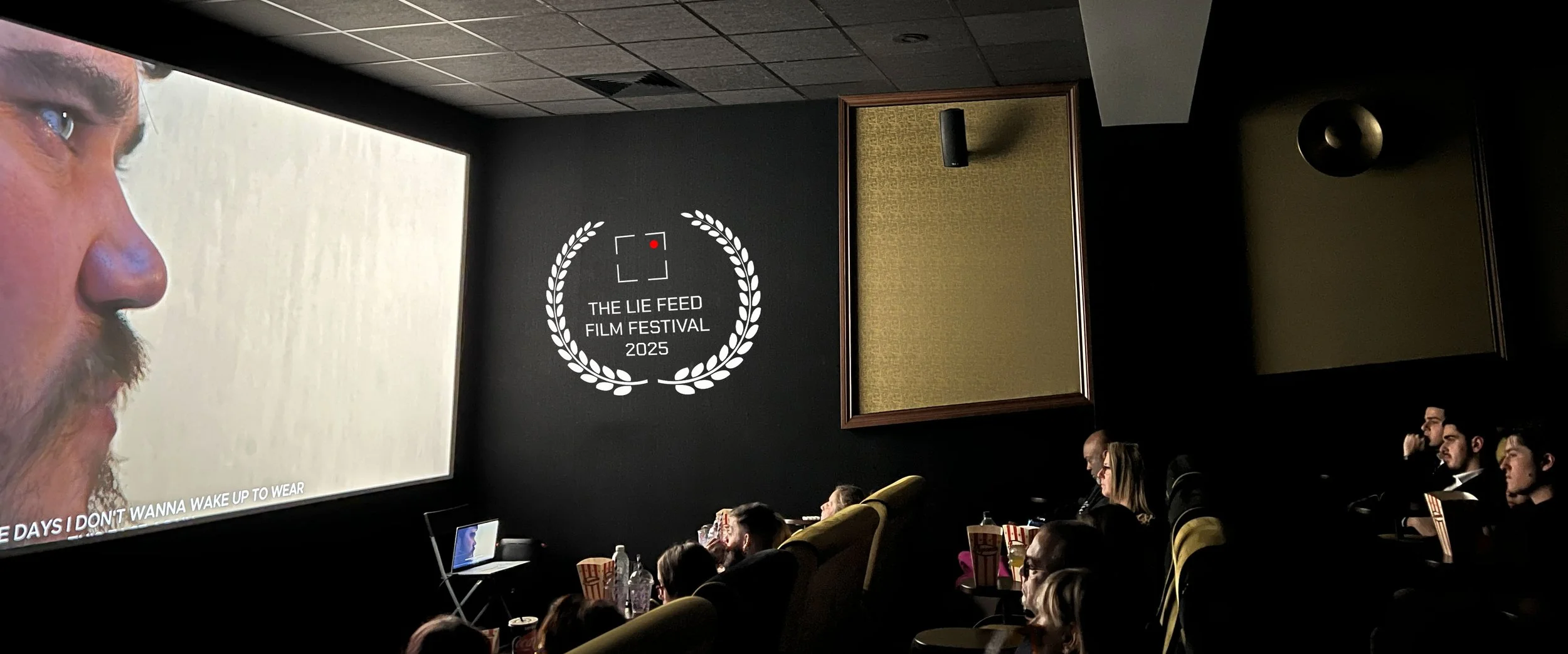 People seated in a darkened movie theater watching a film on a large screen, with a sign on the wall displaying "The Lie Feed Film Festival 2025" and a laurel wreath logo.