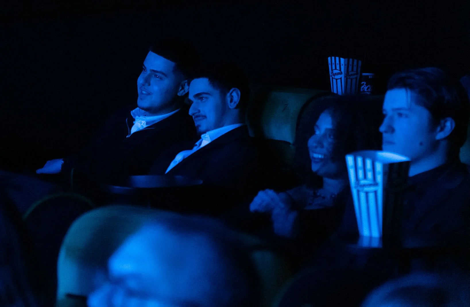 Four people sitting in a dark movie theater, illuminated by blue light, watching a film. Three young men are in the foreground, and a young woman is smiling among them. Popcorn and soda cups are visible on their seats.