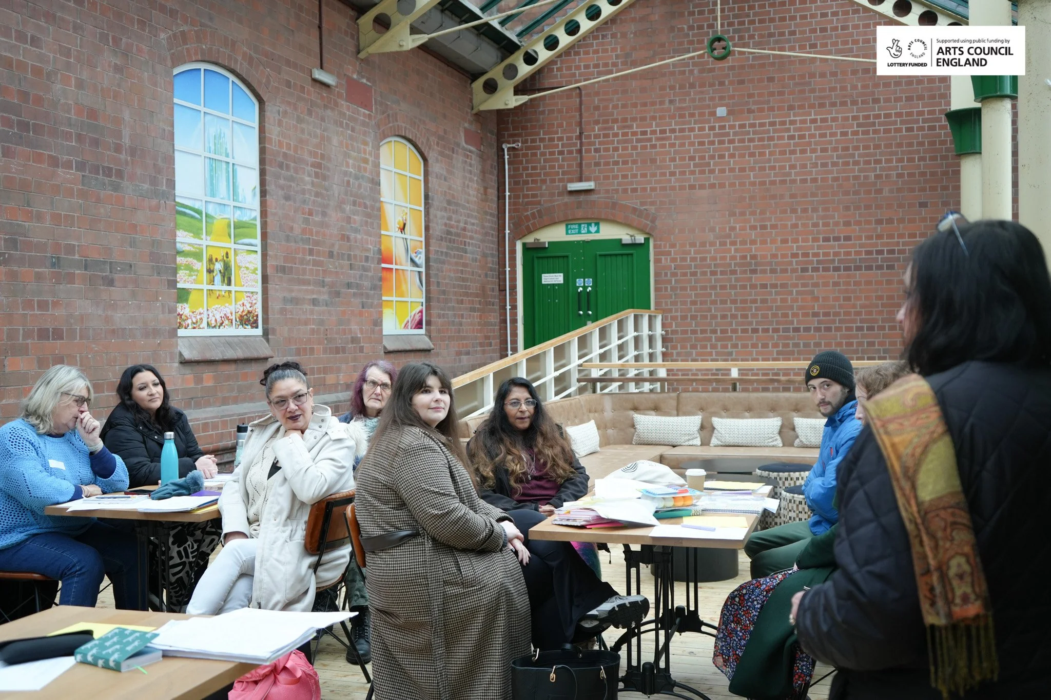 Group of women and one man participating in a meeting or workshop in a spacious, indoor venue with brick walls and large, colorful stained-glass windows. They are seated around tables with papers, notebooks, and a water bottle.
