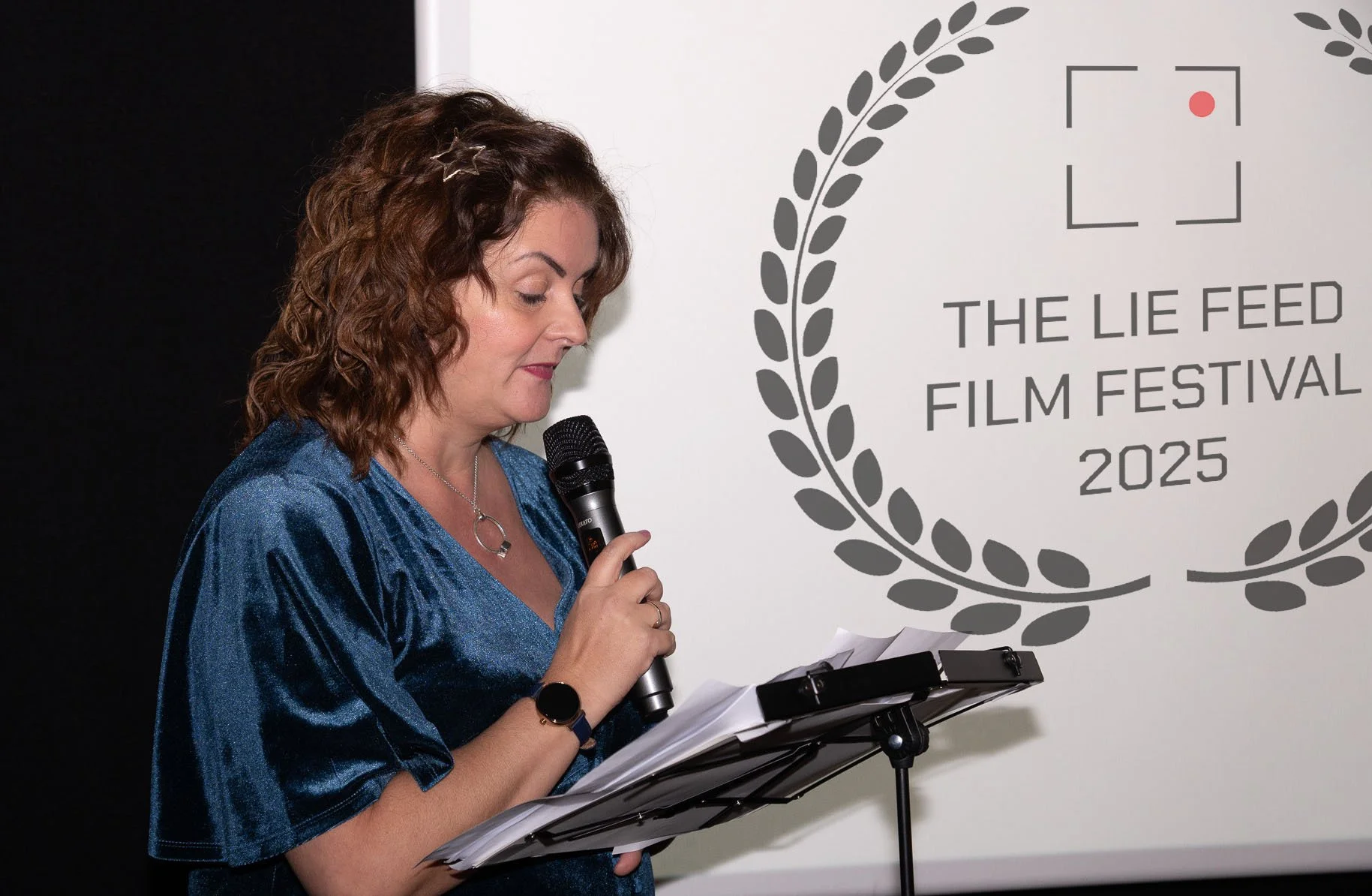 A woman with curly brown hair, wearing a blue velvet dress, speaking into a microphone and reading from papers on a stand in front of a white wall with the logo and text for The Lie Feed Film Festival 2025.