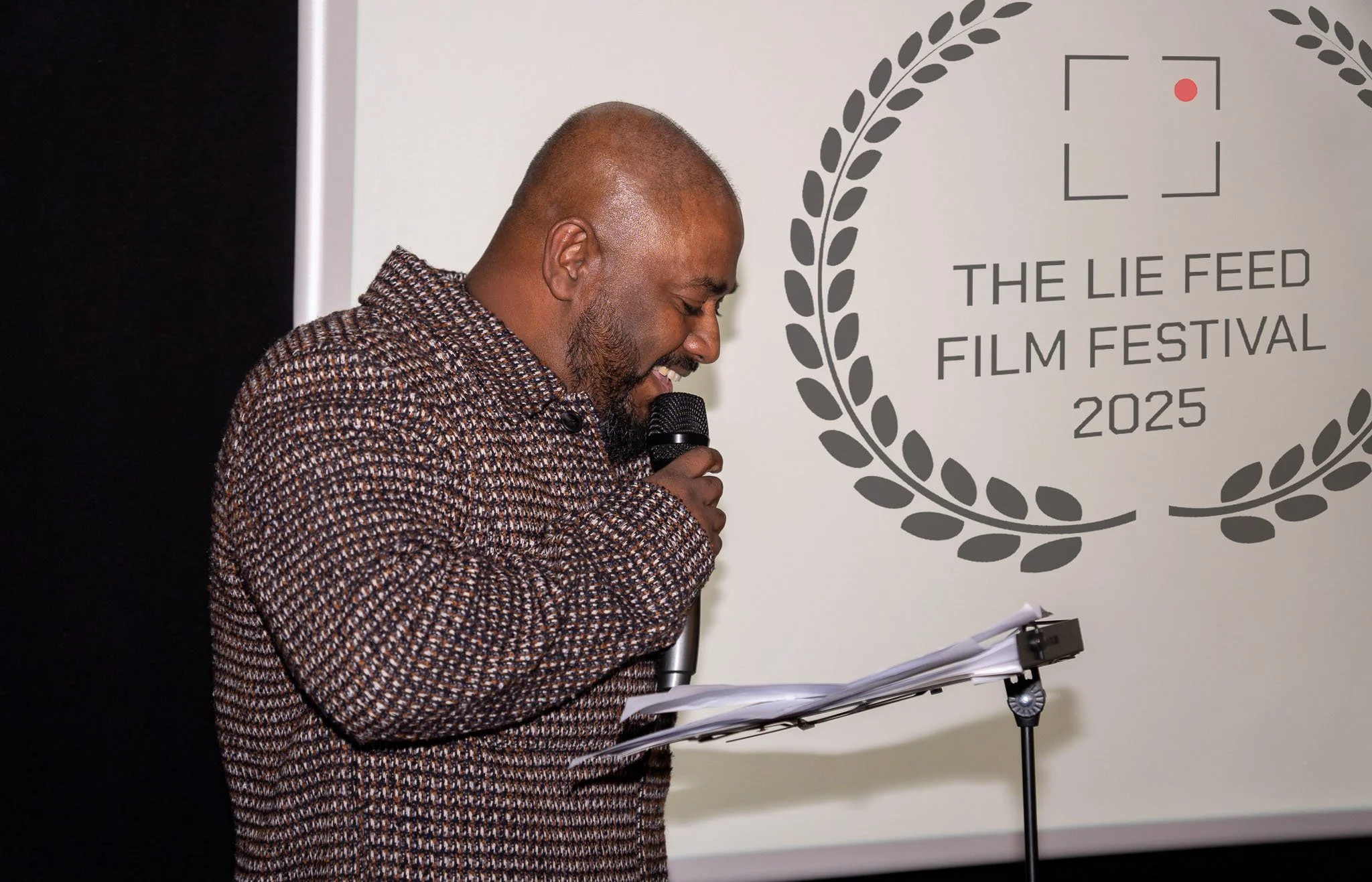 A man with a beard and shaved head speaking into a microphone at a film festival event, with a screen displaying the event's logo in the background.