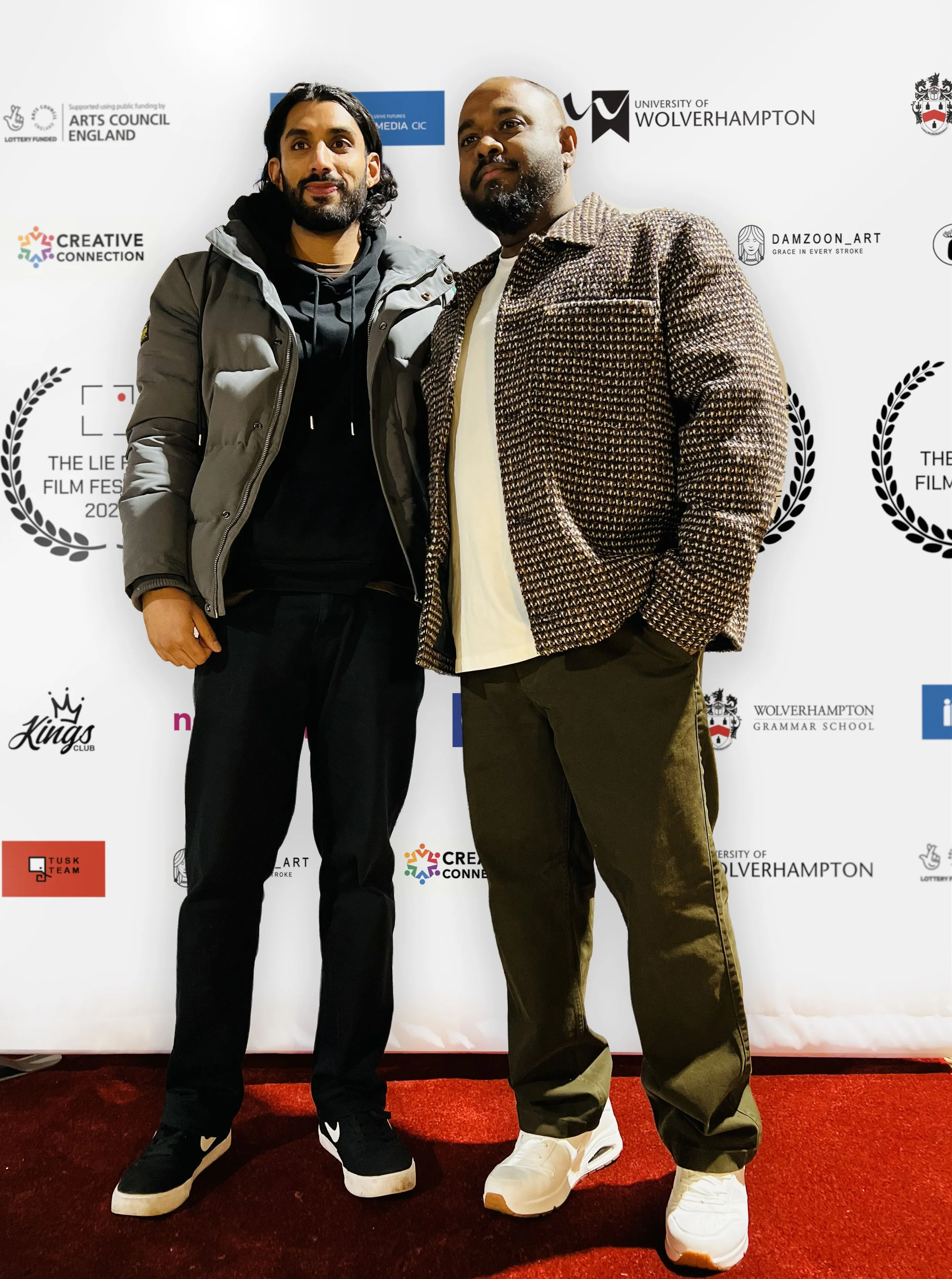 Two men standing side by side, posing in front of a step-and-repeat backdrop at an event. The backdrop displays logos and names of various organizations, including the Arts Council England, Wolverhampton University, and others.