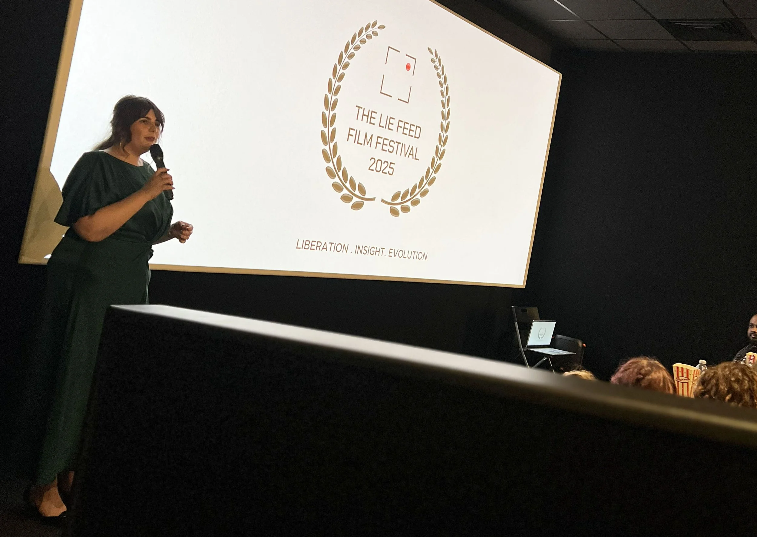 A woman in a dark green dress is speaking into a microphone on stage at the 2023 The Lie Feed Film Festival. Behind her, a large screen displays the festival's logo and the words "Liberation. Insight. Evolution." Audience members are visible in the f