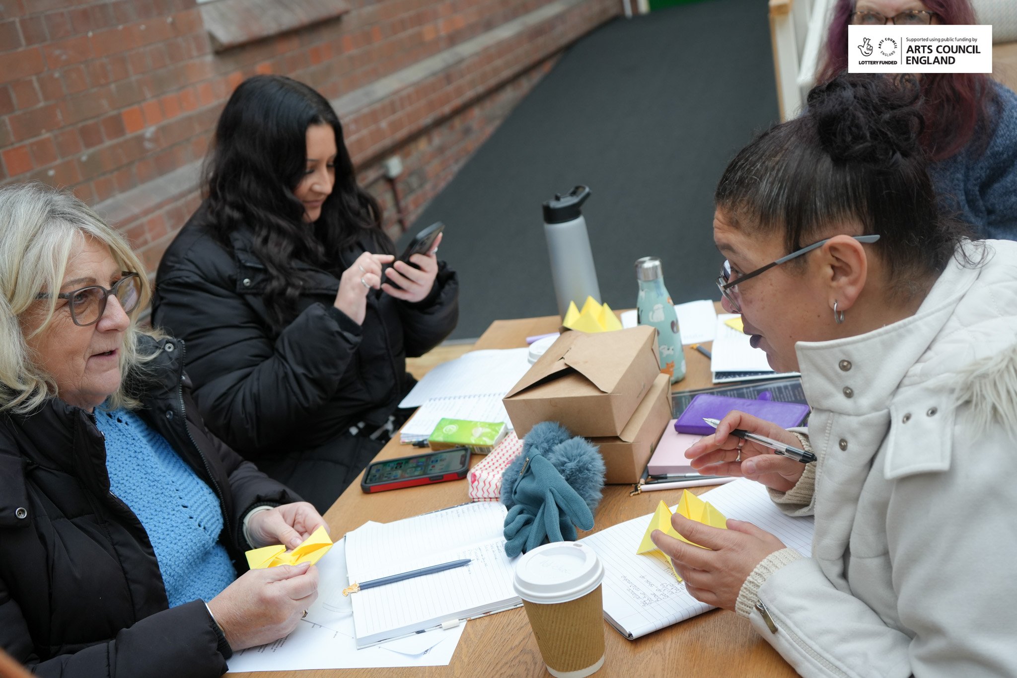 Four women sitting at a table outdoors, engaging in activities with paper crafts, notebooks, and phones, with a brick wall in the background. One woman is holding a yellow paper origami, another is writing, and the others are looking at their phones.