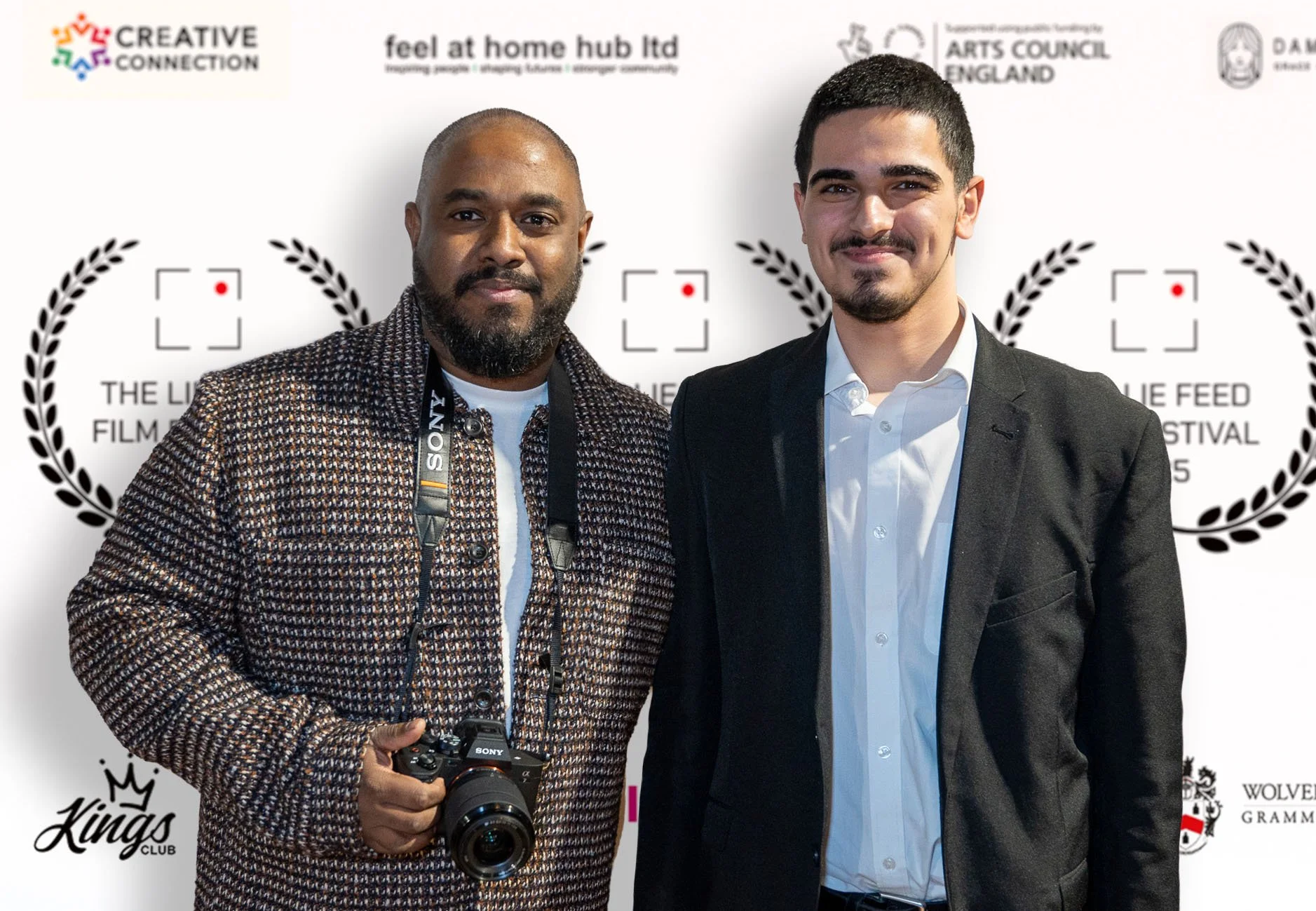 Two men standing side by side at an event with a branded backdrop, one holding a camera, both dressed in business casual attire, smiling at the camera.