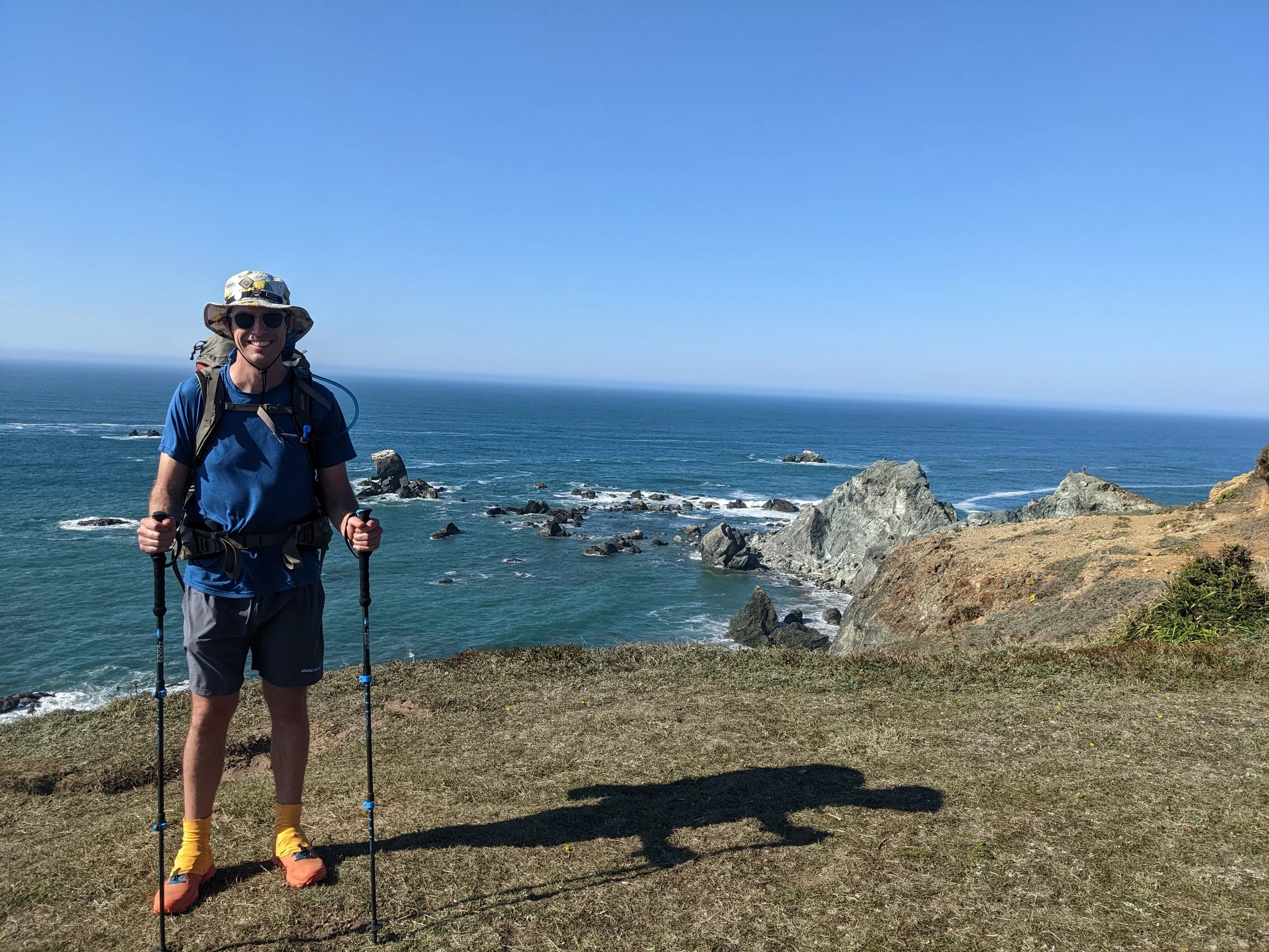 A man in outdoor gear smiling and holding hiking poles stands on a grassy cliff overlooking the ocean with rocks and waves, under a clear blue sky.