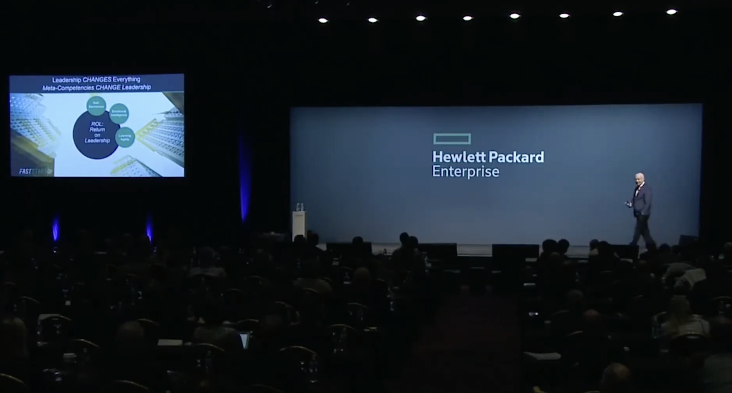 A large presentation stage with a speaker standing next to a giant screen that displays the Hewlett Packard Enterprise logo, while a smaller screen on the left shows slides related to leadership and change.