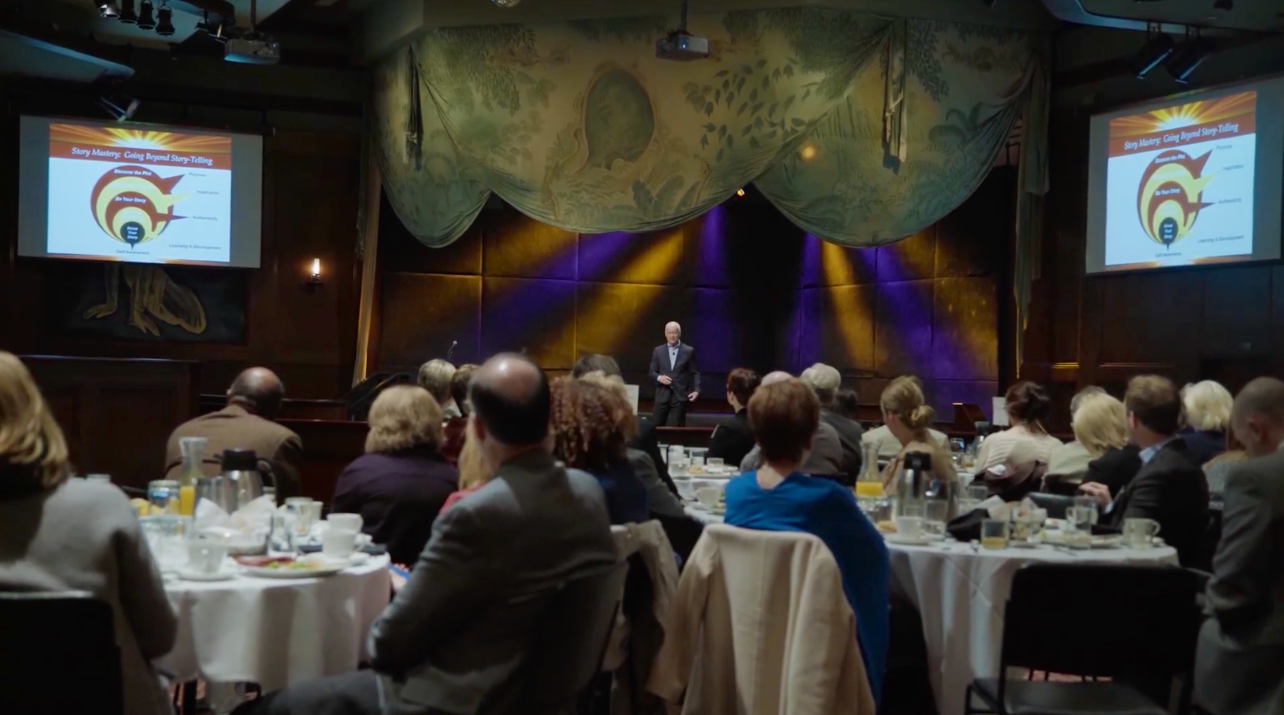A speaker stands on stage in front of an audience in a large, formal dining room, giving a presentation. Two screens display a slide titled 'Story Mastery: Going Beyond Storytelling' with a circular diagram and text. The audience is seated at round tables with breakfast or brunch items.
