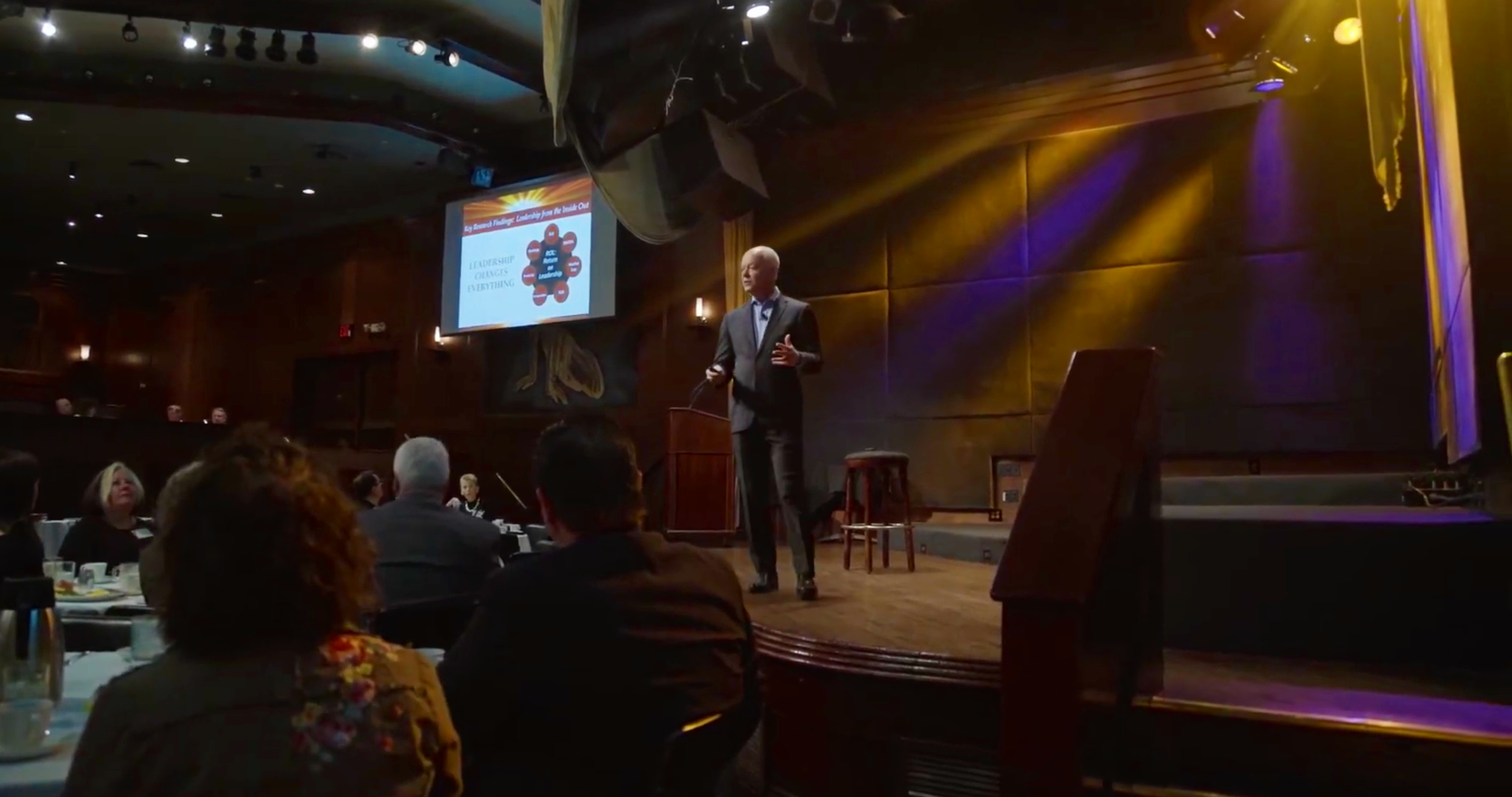 A man in a suit giving a presentation on stage at a conference, with a projected slide behind him, audience seated at tables in a dimly lit room.