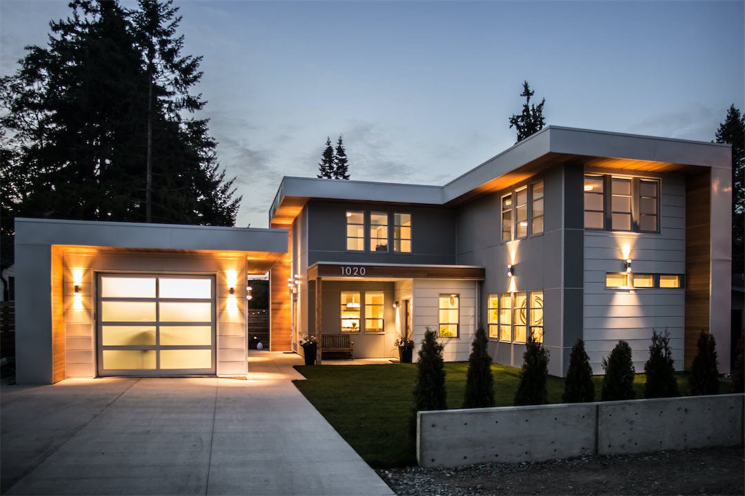 Modern two-story house with a lit garage, sidewalk, and landscaped front yard at dusk.