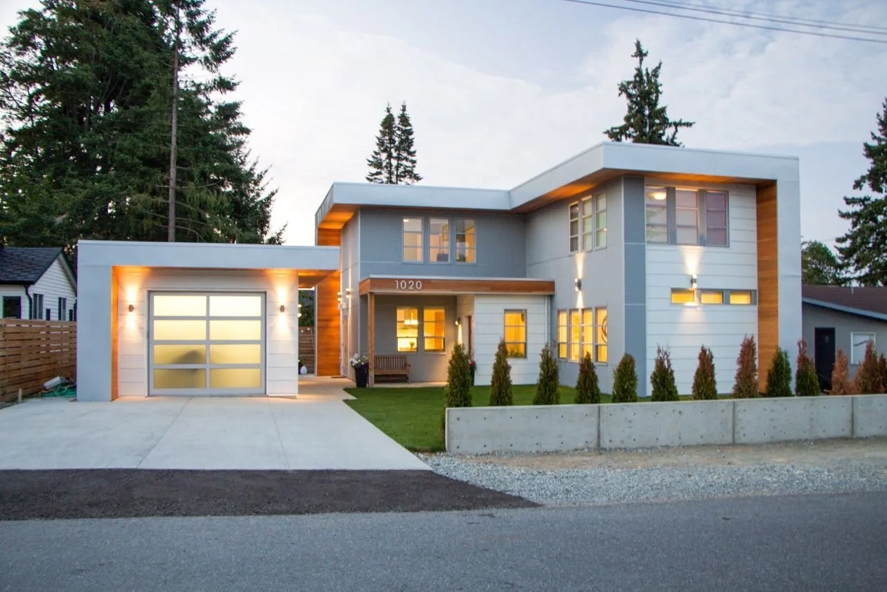 Modern two-story house with a flat roof, large windows, and a combination of white and gray exterior siding, illuminated by outdoor wall lights, with a small front yard and a concrete driveway.