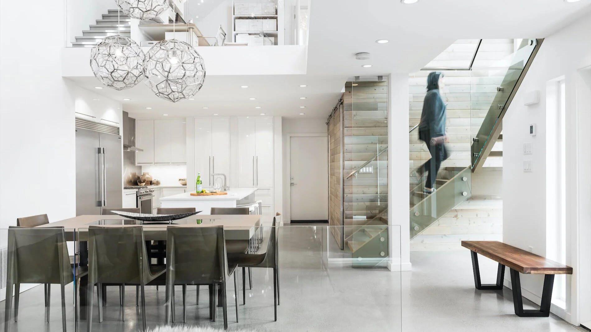 Modern open-concept kitchen and dining area with white cabinetry, stainless steel appliances, a wooden accent wall, a glass staircase with a person walking upstairs, and contemporary lighting fixtures.
