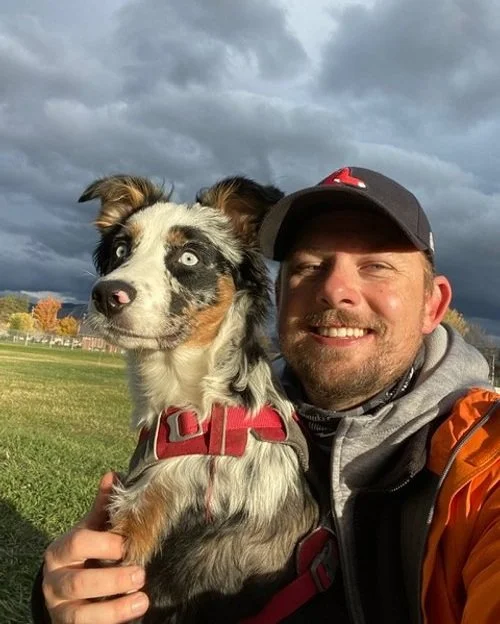 A man smiling with a dog with striking blue eyes outdoors on a cloudy day.