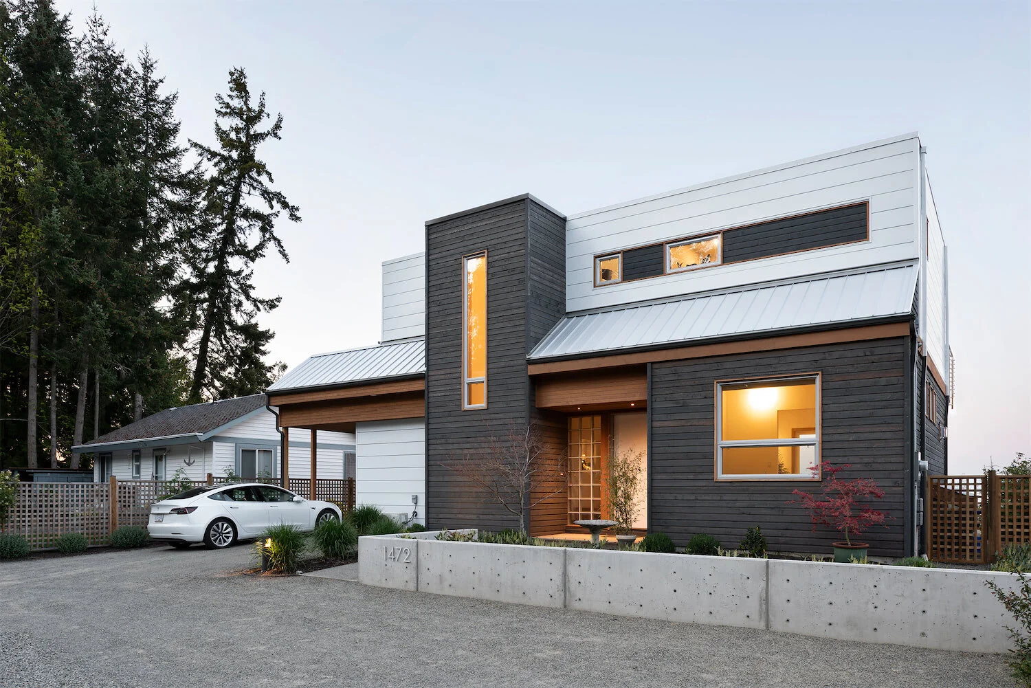 Modern multi-story house with black and white exterior, large windows, and a small front garden with trees and shrubs. A white car is parked in the driveway.