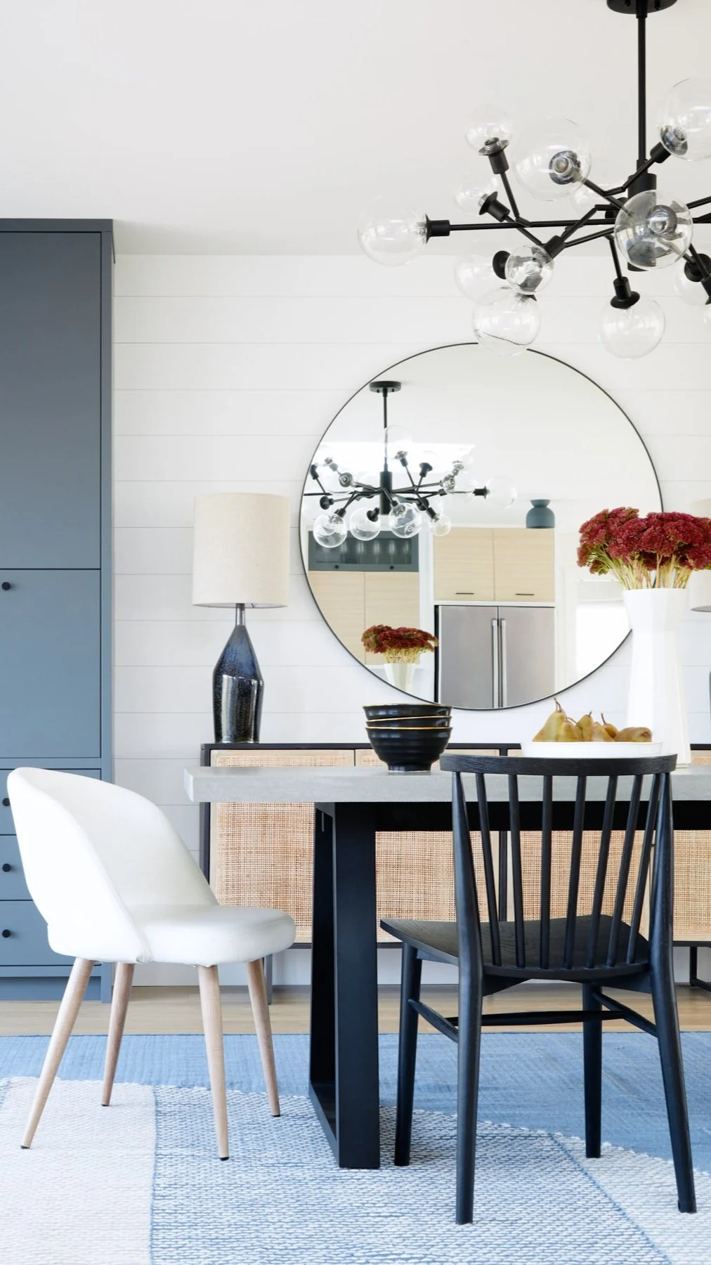 A modern dining room with a black table, white and black chairs, a large round mirror, a black chandelier with multiple glass globes, a tall black lamp with a white shade, and a vase with red flowers.