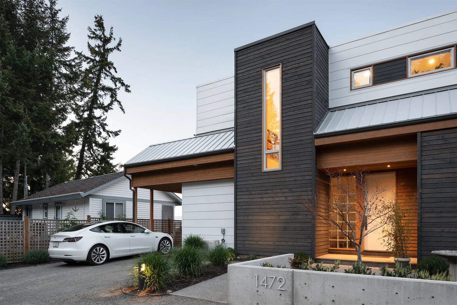 Modern house with black and white exterior, metal roof, and tall narrow window. A white car is parked in the driveway, and a small yard with plants and trees is visible. The house number 1472 is displayed on a concrete wall.