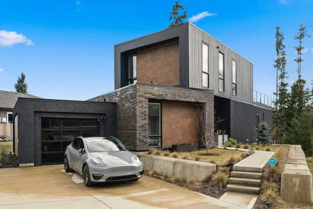 Modern two-story house with a gray and brick exterior, large windows, and a concrete driveway with a silver Tesla parked in front.