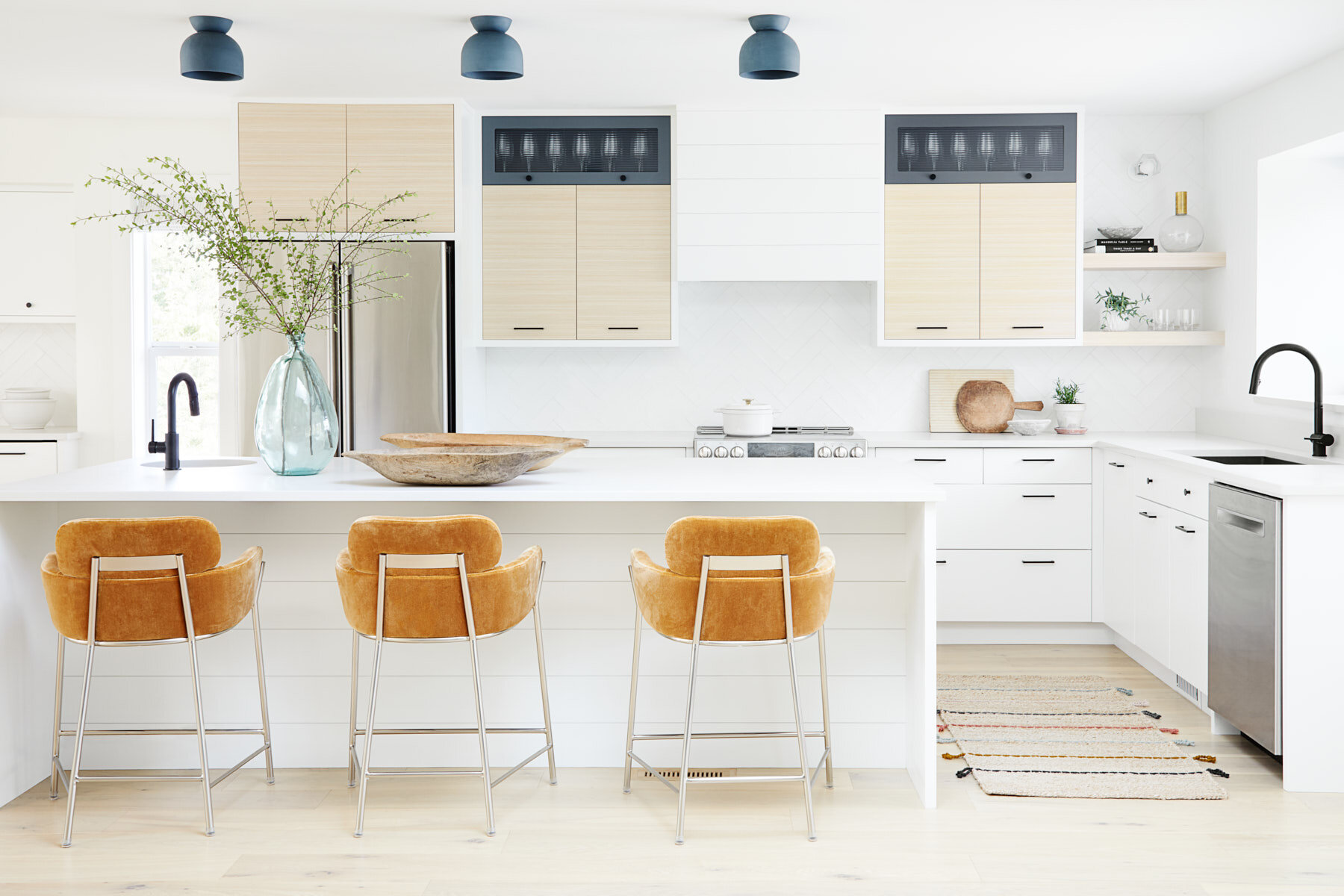 Modern white kitchen with beige cabinets, black fixtures, kitchen island with three tan barstools, decorative vase with greenery, and various kitchen items.