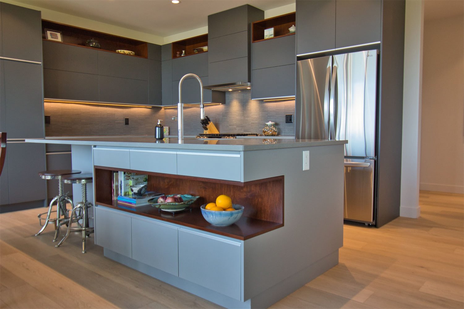 Modern kitchen with gray and wood cabinetry, stainless steel refrigerator, and a kitchen island with a sink, bowl of lemons, and magazines.