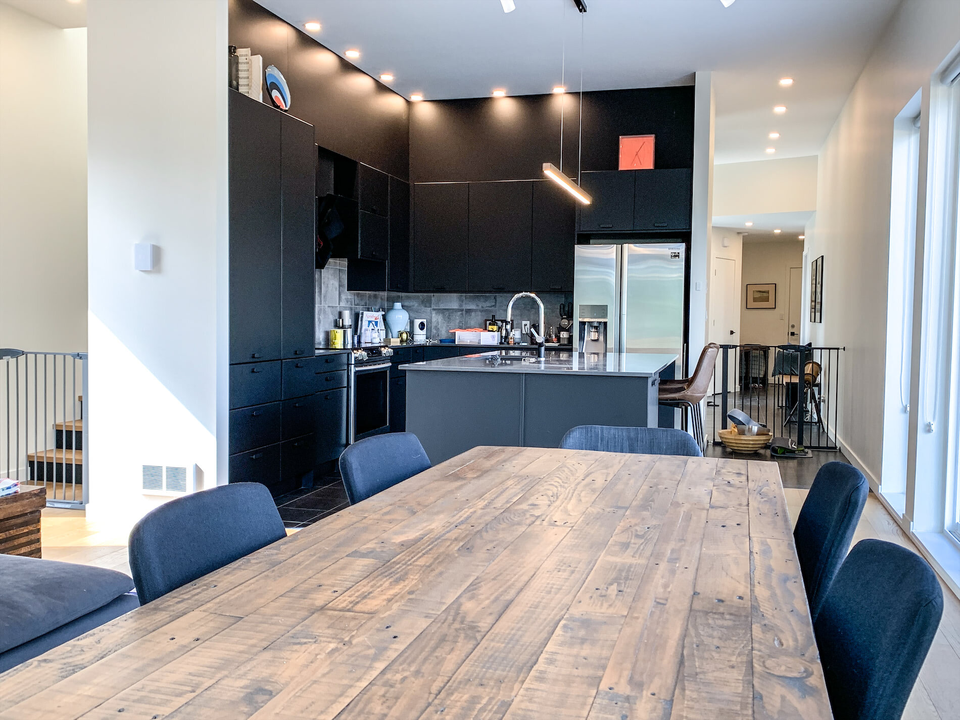 Modern kitchen with black cabinets, stainless steel appliances, and a kitchen island with a sink. There is a dining table with chairs in the foreground. The space is well-lit with ceiling lights and natural light from large windows.