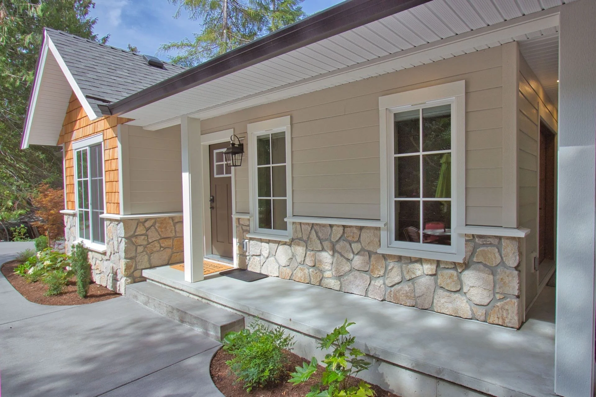 Front view of a house with beige siding, stone foundation, white window frames, and a brown front door, surrounded by a small garden and planted plants.