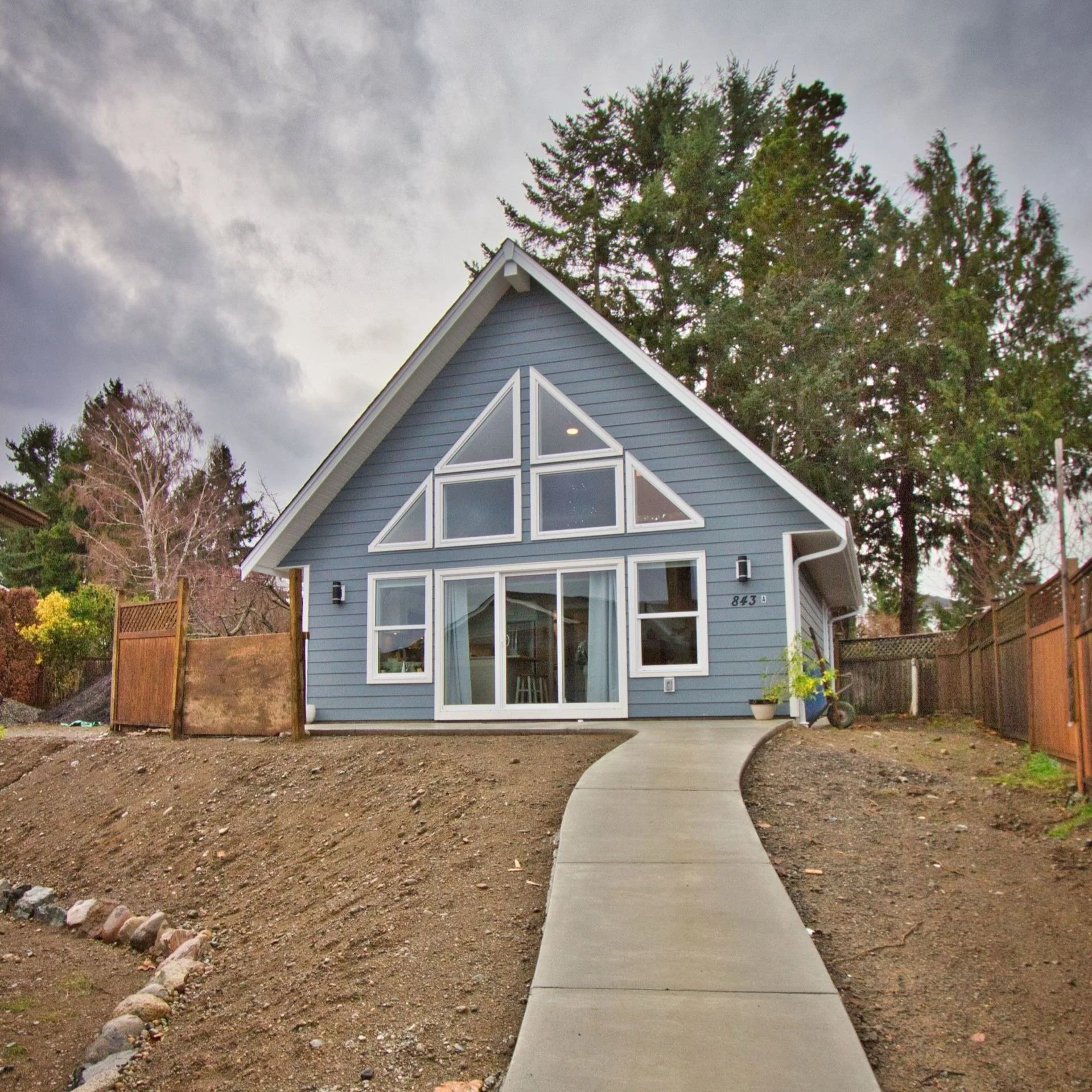 A house with blue siding and white trim, featuring large triangular windows on the upper floor, with a curved concrete pathway in the front yard, and surrounded by trees and fencing under a cloudy sky.