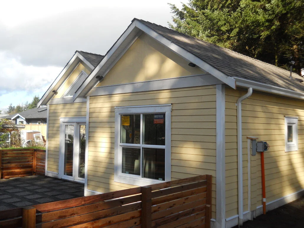 Yellow house with white trim, double glass doors, and a patio area with a wooden fence. The house has a sloped roof with gray shingles and appears to be in a suburban neighborhood.