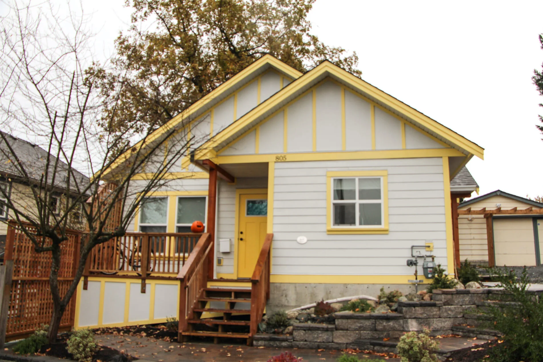 A yellow and gray house with a small front porch, stairs, and a pumpkin on the porch, surrounded by trees, shrubs, and a stone retaining wall in a residential neighborhood.