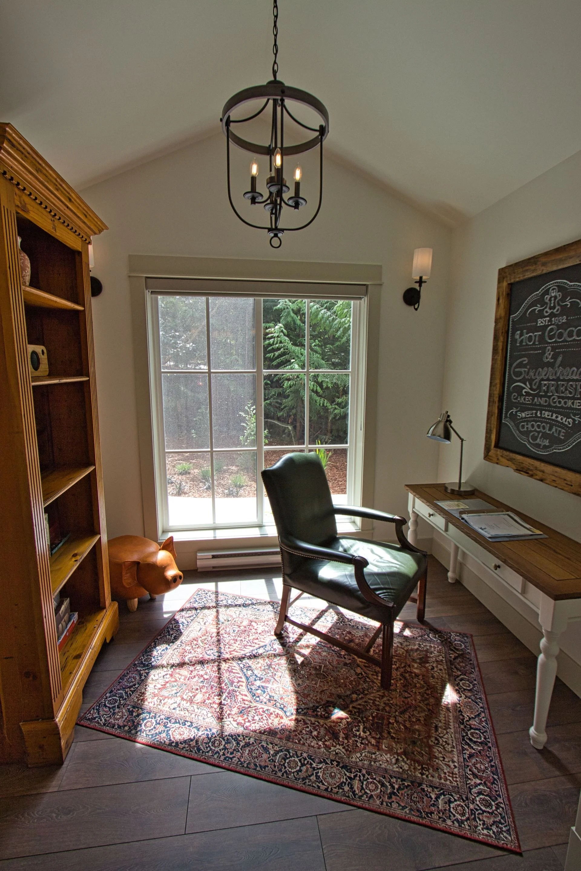 A cozy home office with a large window showing trees outside. There's a black leather armchair, a wooden desk with a lamp, a framed chalkboard sign on the wall, a wooden bookshelf, and a pig-shaped stool. An ornate rug covers part of the dark wood floor, and a modern chandelier hangs from the ceiling.