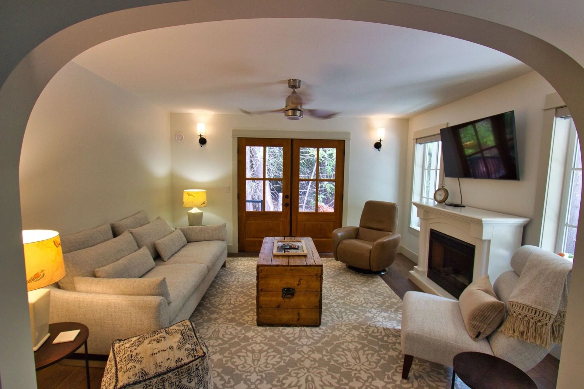 Living room with beige sofas, a wooden coffee table, a beige armchair, a fireplace, a wall-mounted TV, two lamps, and large windows showing trees outside.