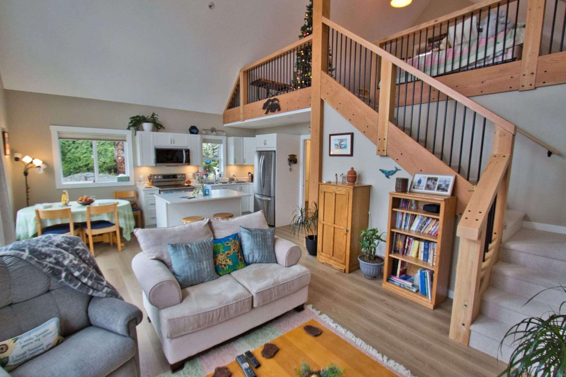 Living room with beige sofa, armchair, and pillows, open kitchen with white cabinets and a dining table, staircase with wooden railing, decorated for Christmas with a Christmas tree and star-shaped decorations.