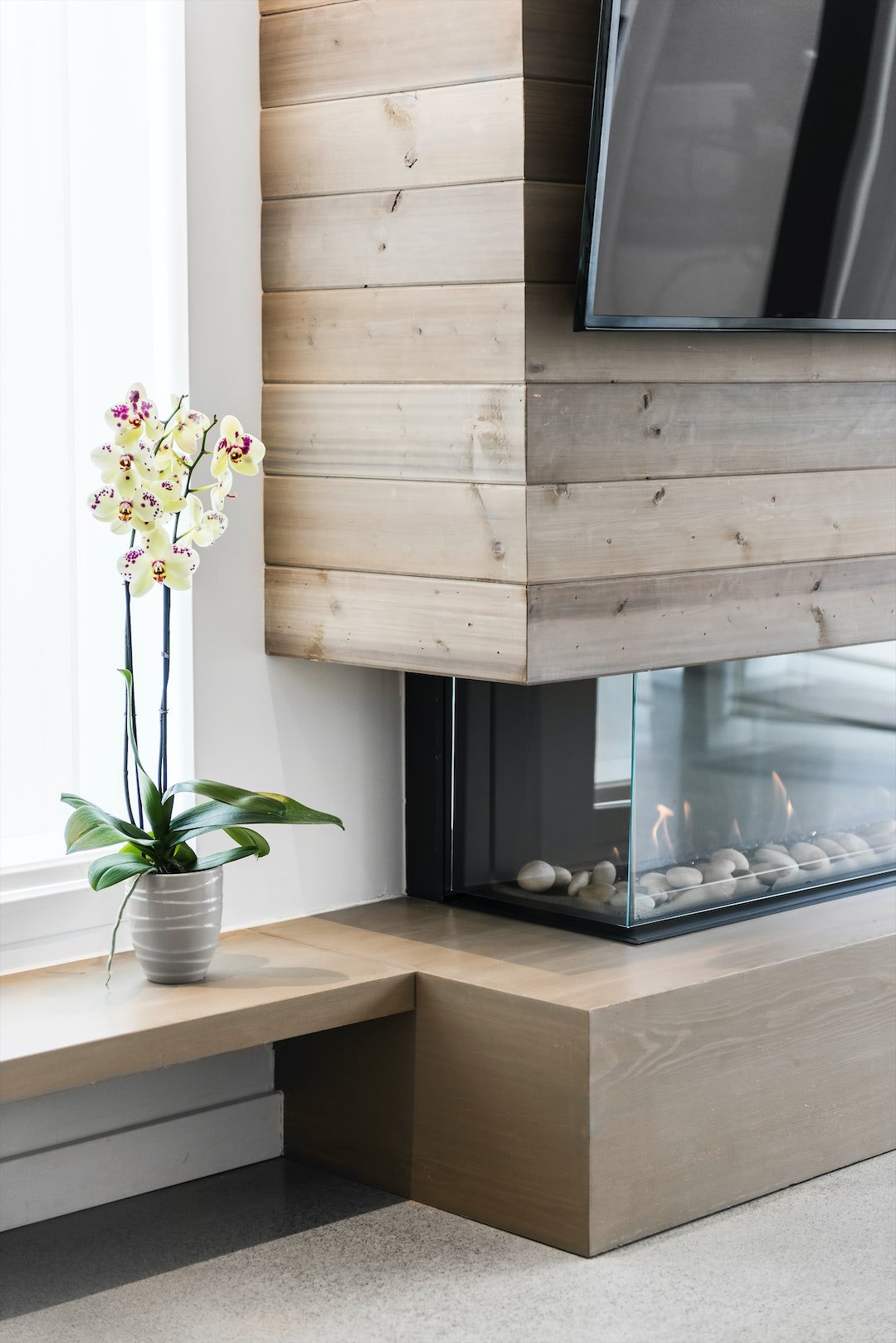 A decorative interior space featuring a white orchid plant in a striped pot on a wooden ledge, a corner of a flat-screen TV, a modern fireplace with glass panels and white stones, and a wooden wall with horizontal planks.