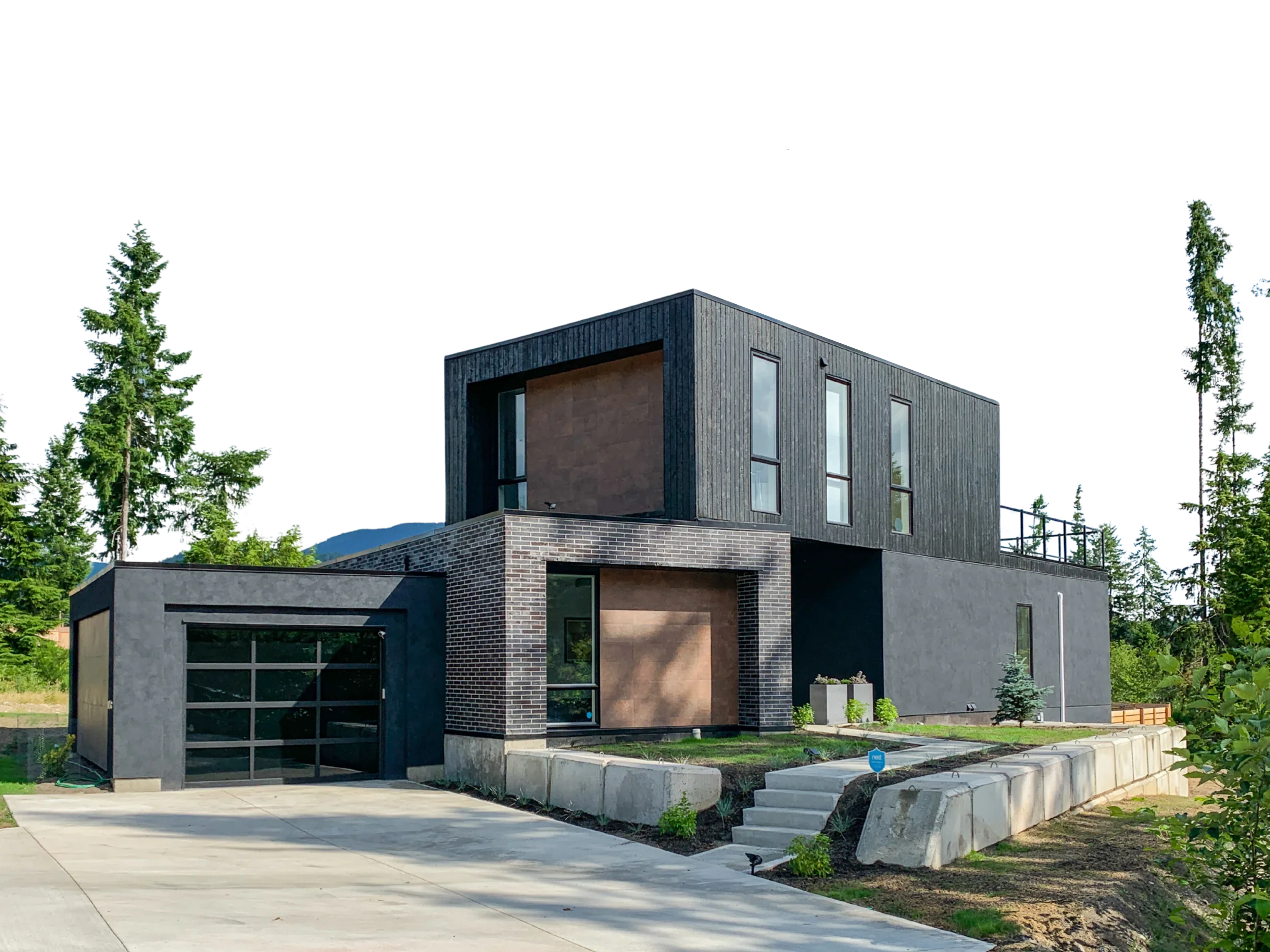 Modern two-story house with a flat roof, dark vertical siding, large windows, and a mix of brick and wood exterior walls, surrounded by trees and a paved driveway.