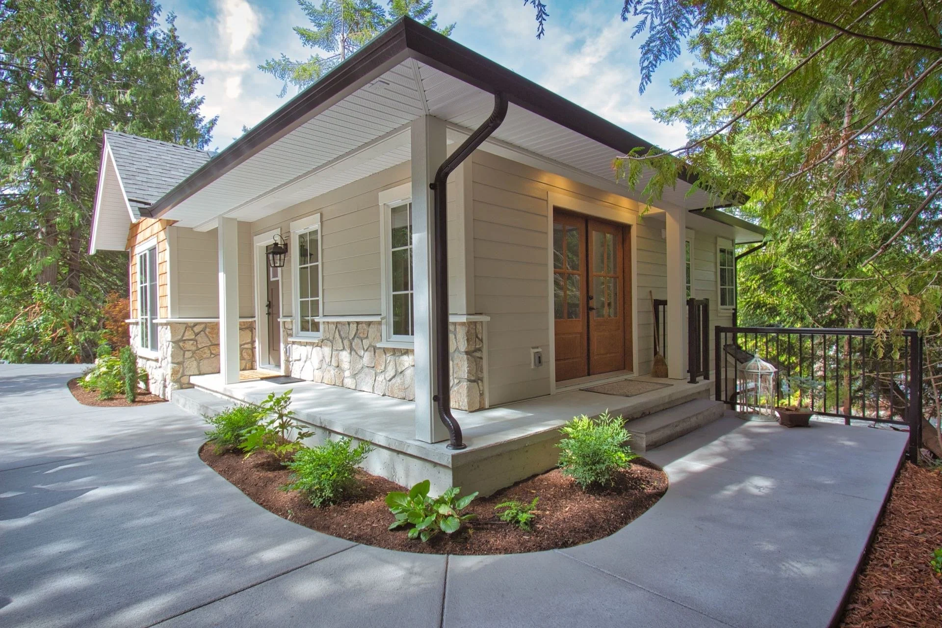 Front of a beige house with stone accents near the entrance, surrounded by lush green trees, with concrete steps leading to the wooden front door. A black railing surrounds a small porch area with decorative items including a birdcage. The sky is partly cloudy with patches of blue.
