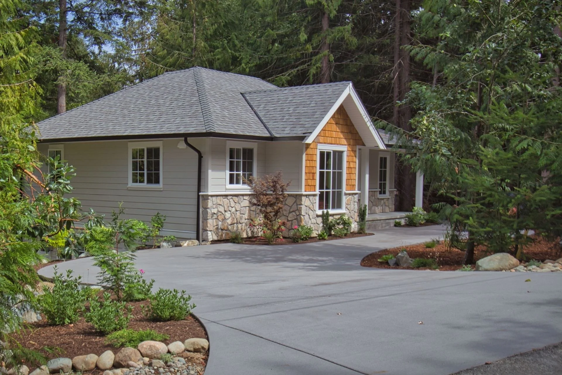 A house with a gray shingle roof, white siding, and large windows, surrounded by green trees and shrubs, with a curved concrete driveway in front.
